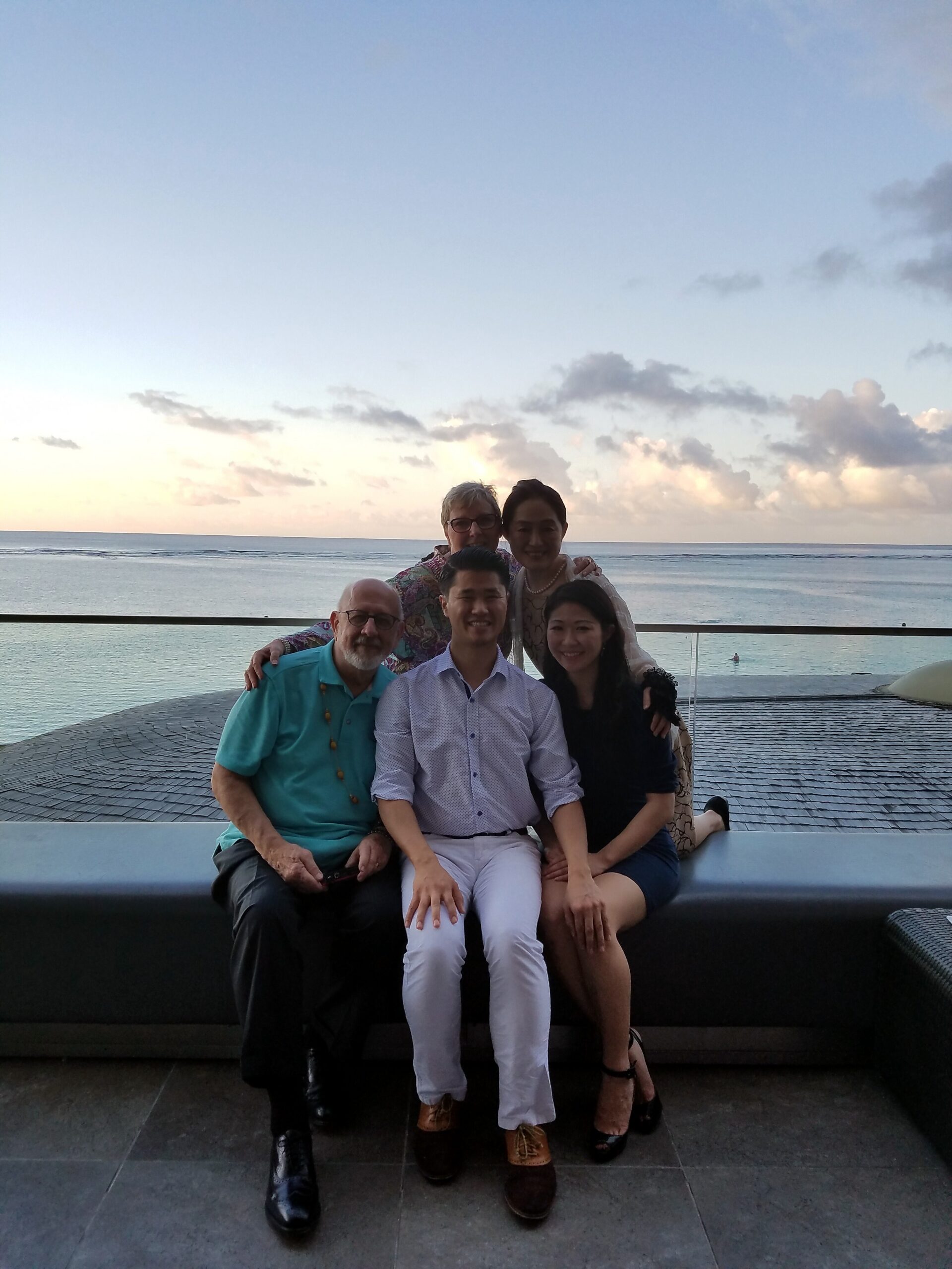 Group of five people posing together on a seaside bench, with ocean and pastel sunset sky in the background.