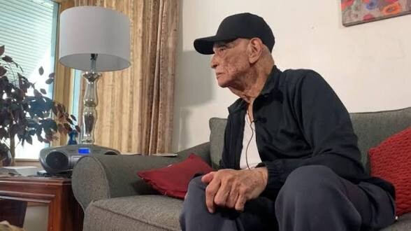 Older man in a black cap and jacket sits on a gray sofa in a living room, looking to the right.
