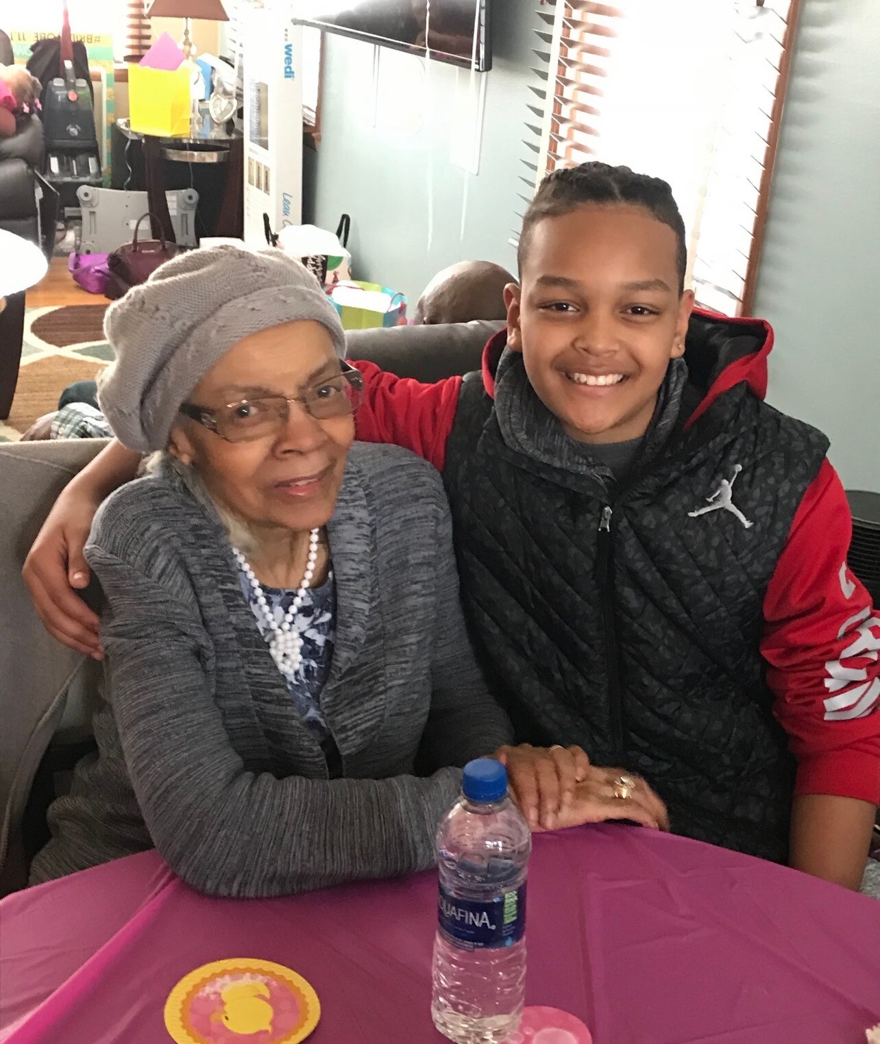 Young man with arm around an elderly woman smiling at a family gathering, seated at a pink table with a water bottle nearby.