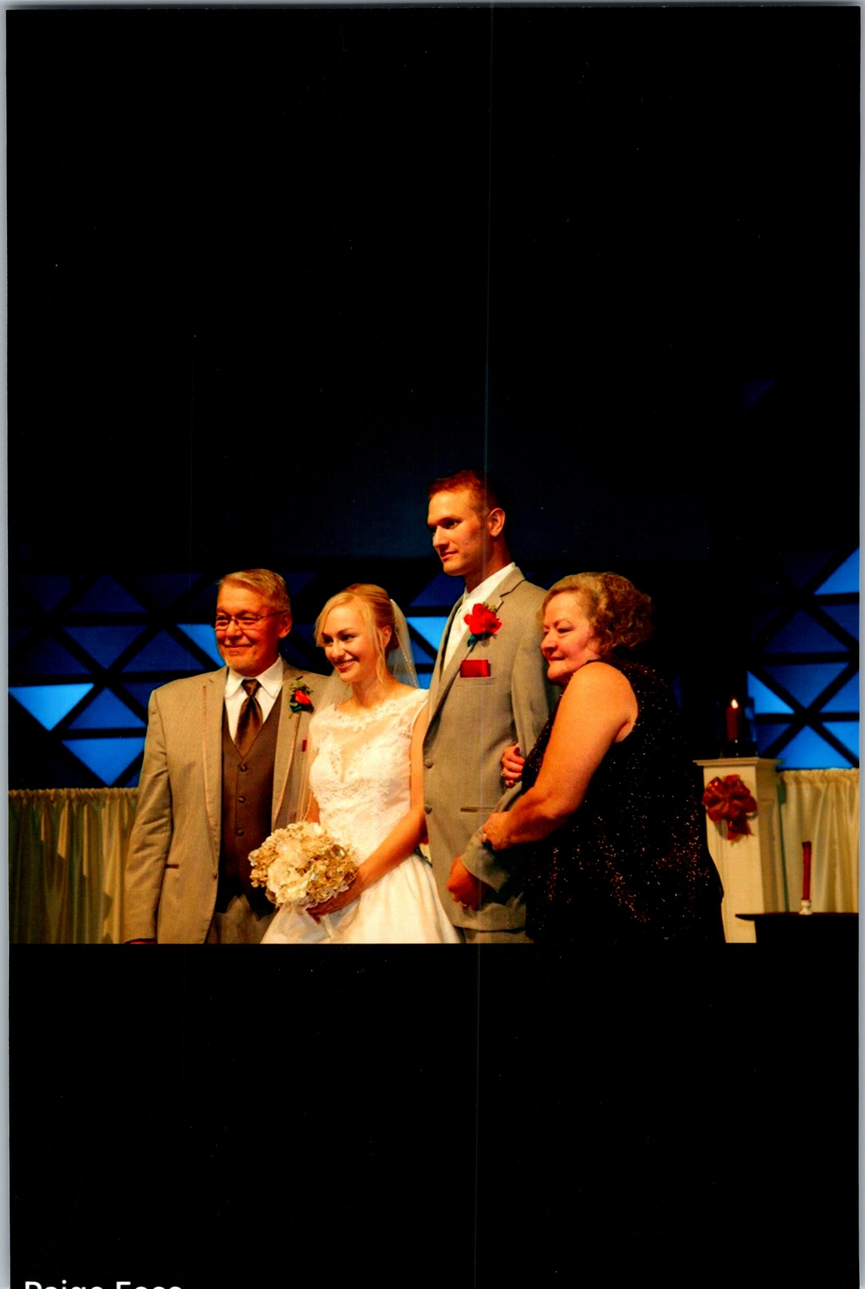 Bride and groom stand with two older relatives on a stage, smiling for a wedding photo; the bride holds a bouquet and wears a white gown, the groom wears a light gray suit with a red boutonniere, and an older man in a tan suit while an older woman in a black dress stands beside them.