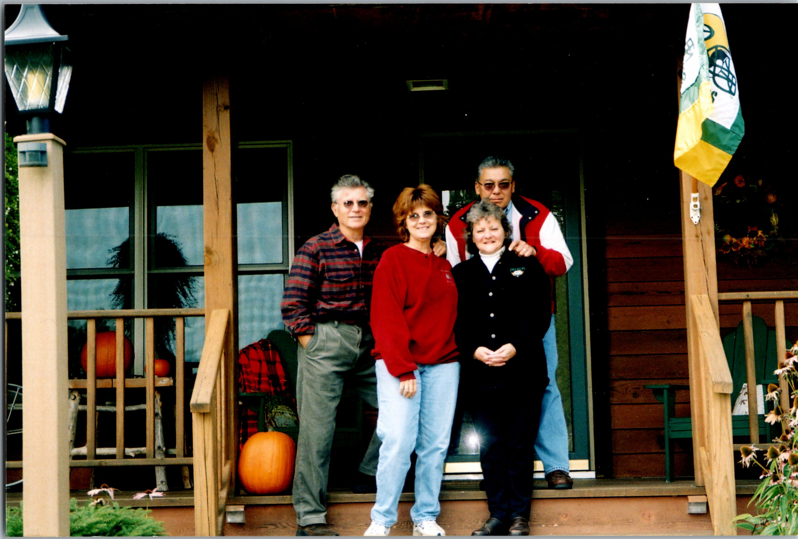 Four adults posing on a wooden porch with pumpkins and autumn decor in front of a house.