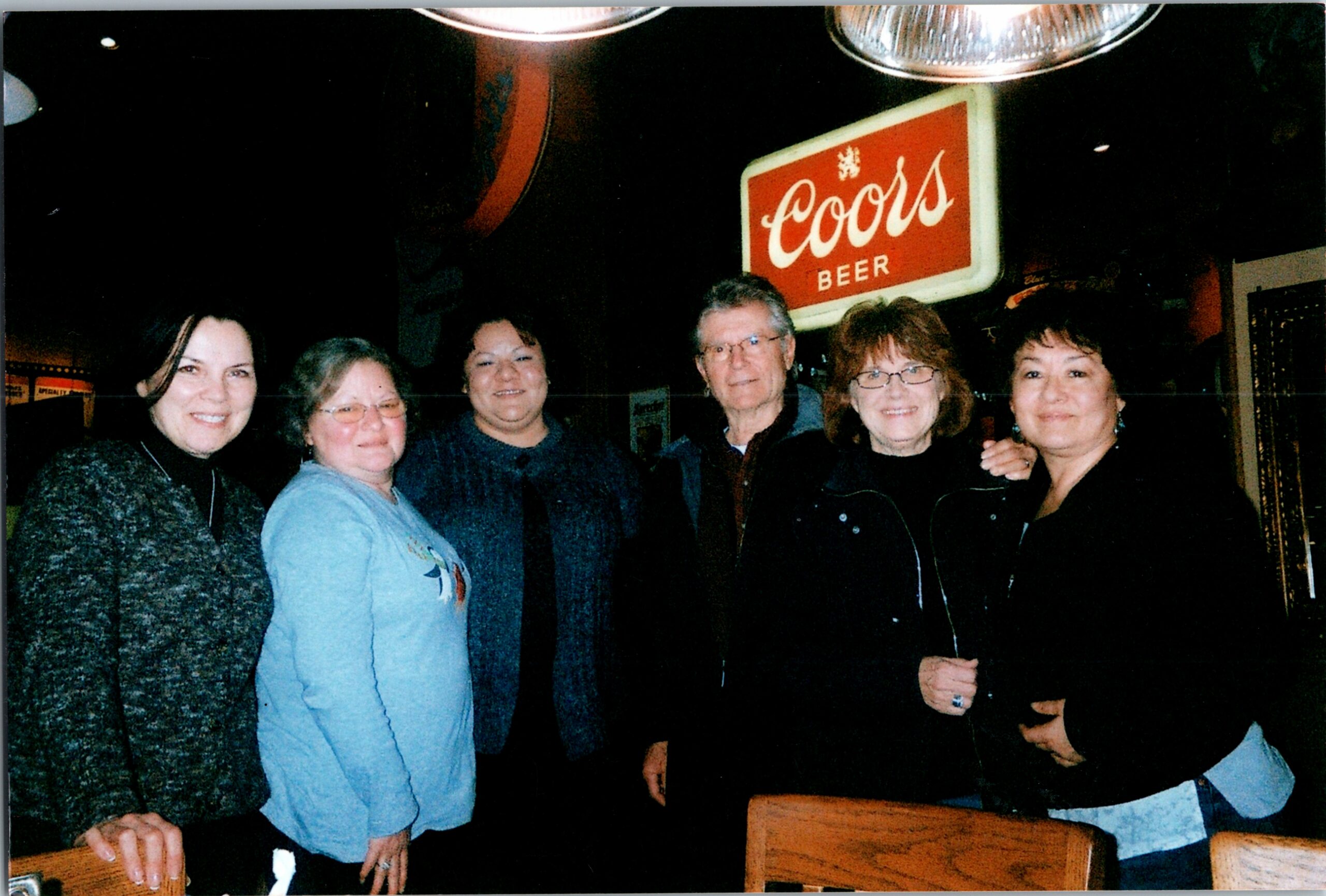 Six adults posing together in a dim venue with a Coors Beer sign in the background, smiling at the camera.