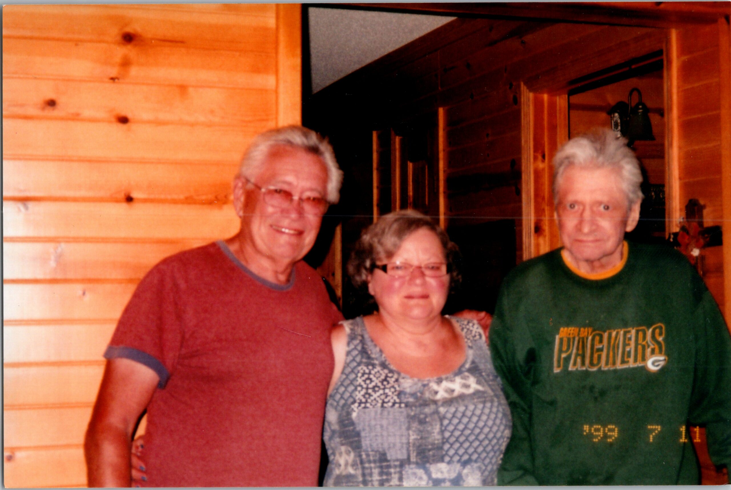 Three smiling seniors posing together indoors in a wood-paneled room; left man in a red shirt, center woman in a patterned blouse, right man in a green Packers shirt.