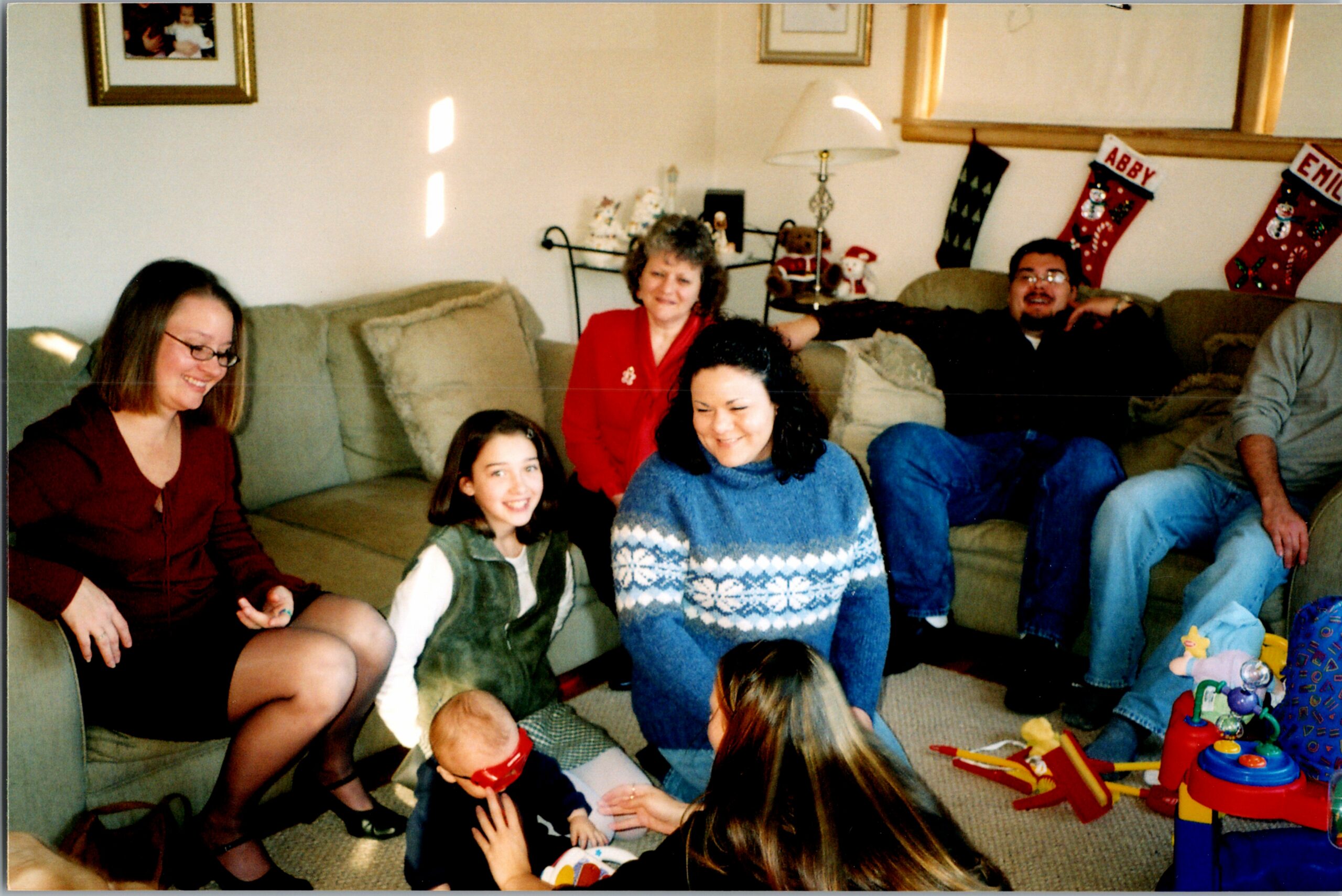 Family gathered on a sofa and floor in a living room during Christmas, with stockings hanging on the wall in the background.