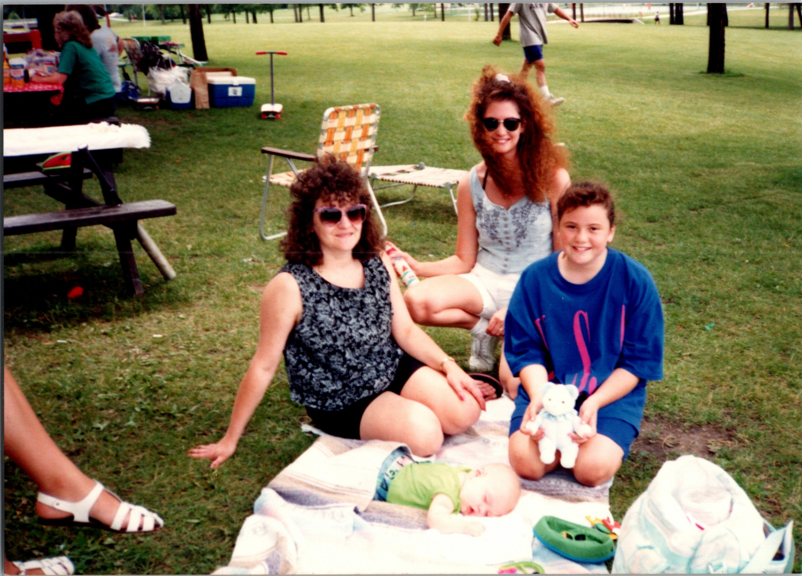 Group of people at a park picnic on a sunny day, sitting on blankets with toys and snacks spread on the grass.