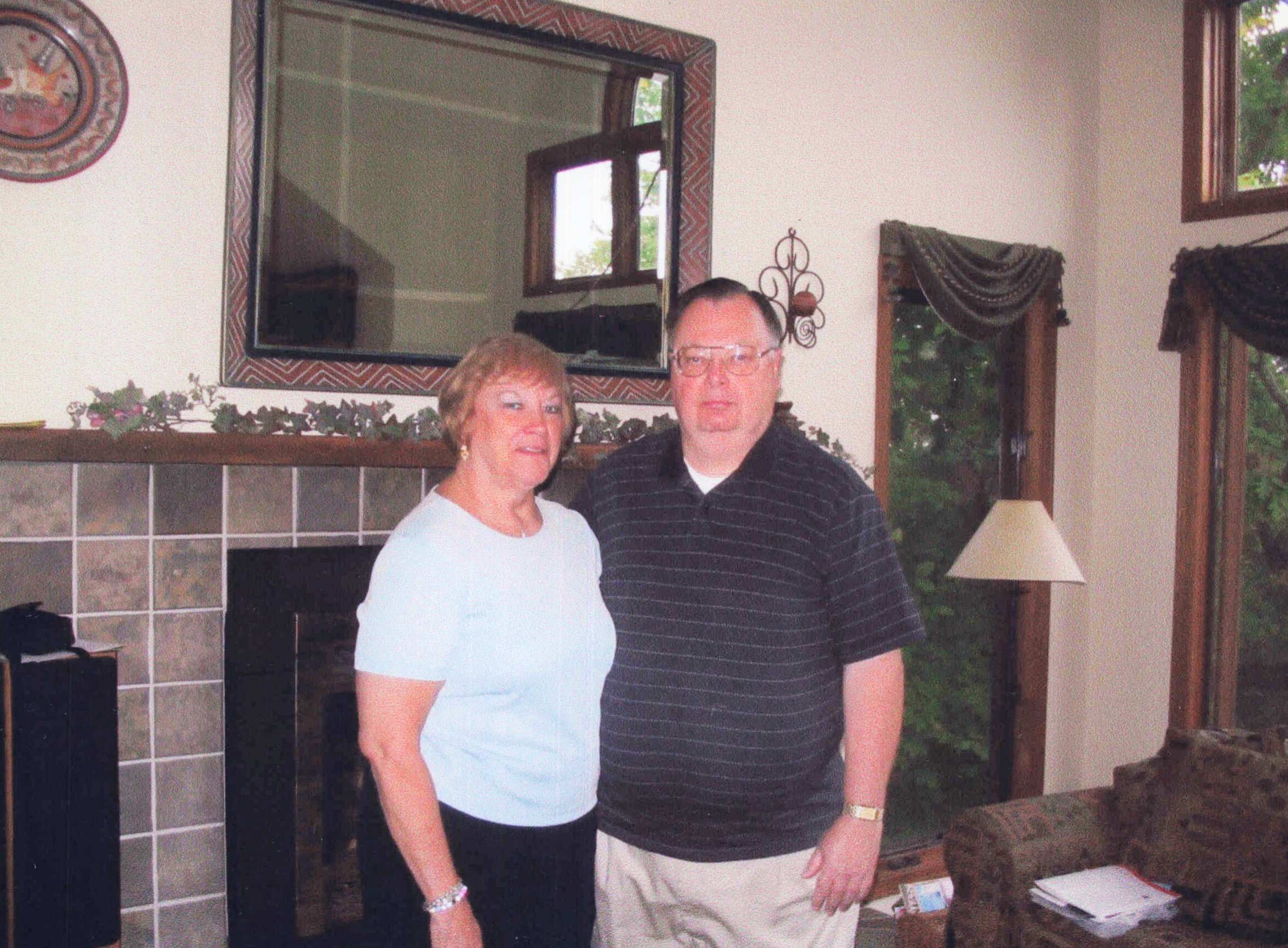Two adults posing together in a living room, standing in front of a fireplace and large mirror.