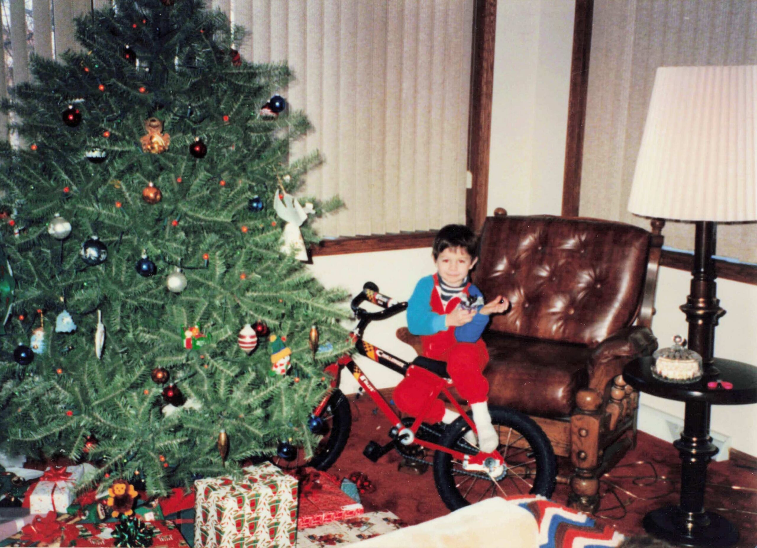 Young boy in red and blue pajamas sits on a small red bicycle beside a decorated Christmas tree with presents underneath.