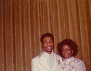 Couple posing in front of a gold curtain; man in a white suit and woman in a patterned dress.
