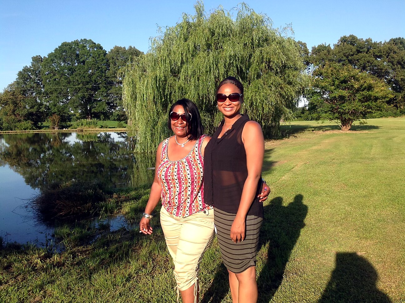 Two women standing together by a pond in a sunny park, both wearing sunglasses and summer outfits.