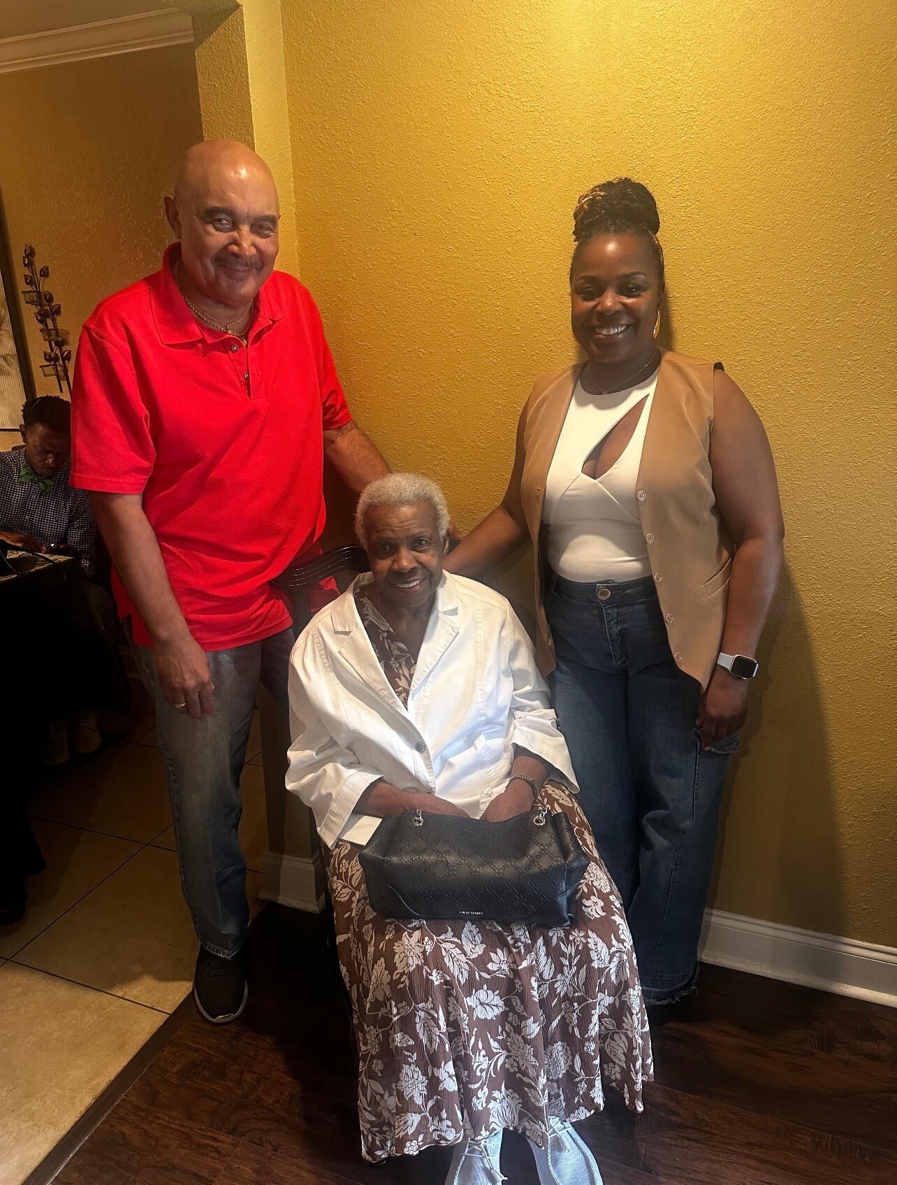 Three people pose together indoors: an elderly woman seated in front wearing a white blazer and a floral skirt, flanked by a man in a red polo shirt and a smiling woman in a beige vest and white top against a yellow wall.