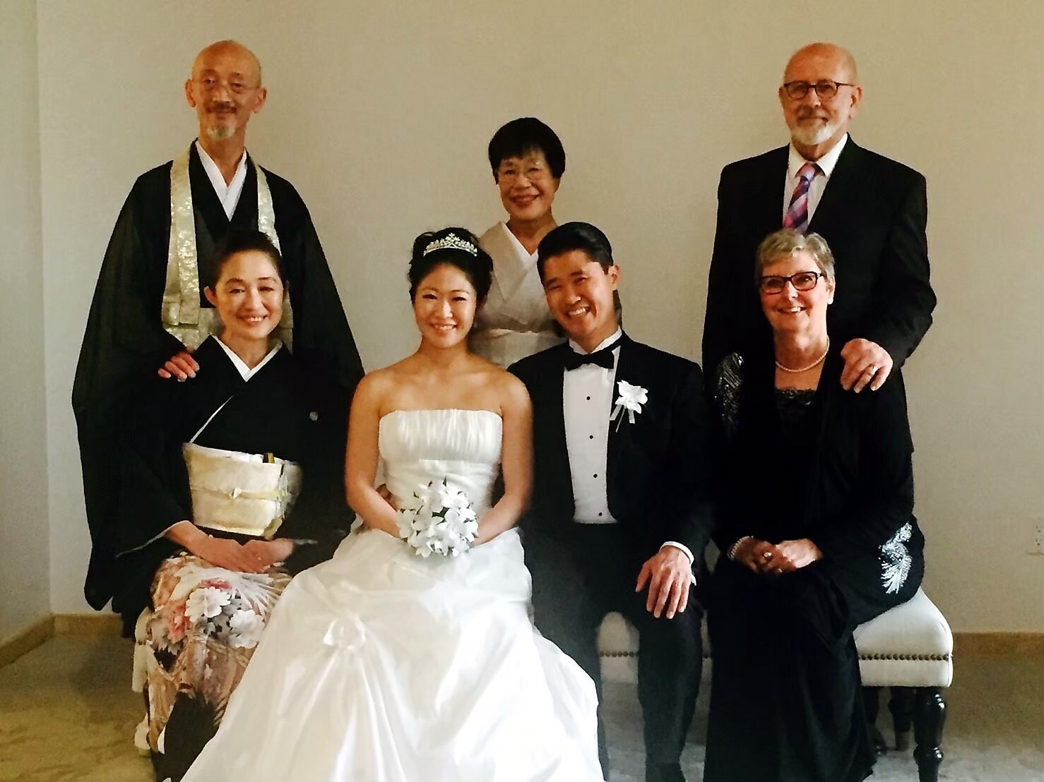 Wedding group portrait: bride in white strapless gown holding a bouquet, groom in black tuxedo beside her, seated with family and officiants behind.