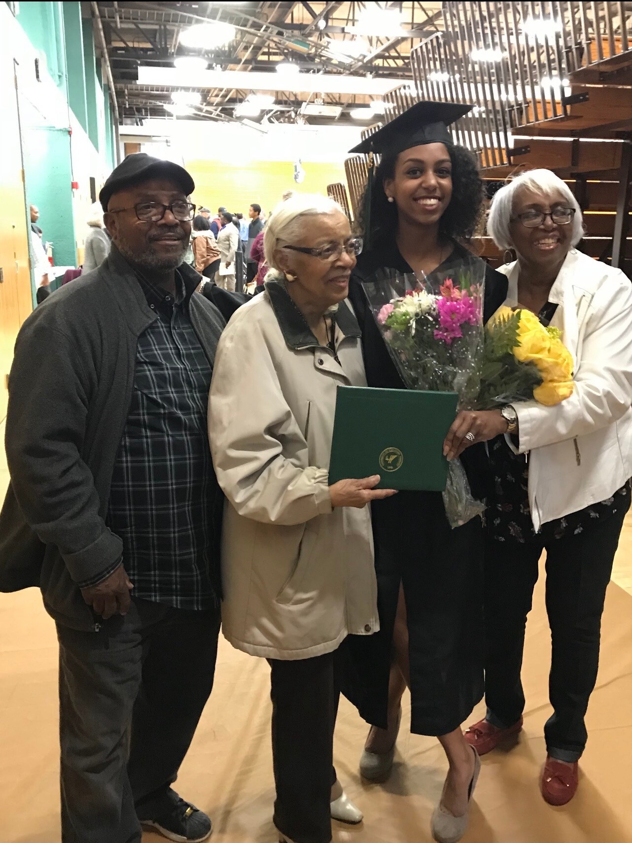 Graduating student in cap and gown holding flowers and a diploma, flanked by smiling family indoors at a ceremony.