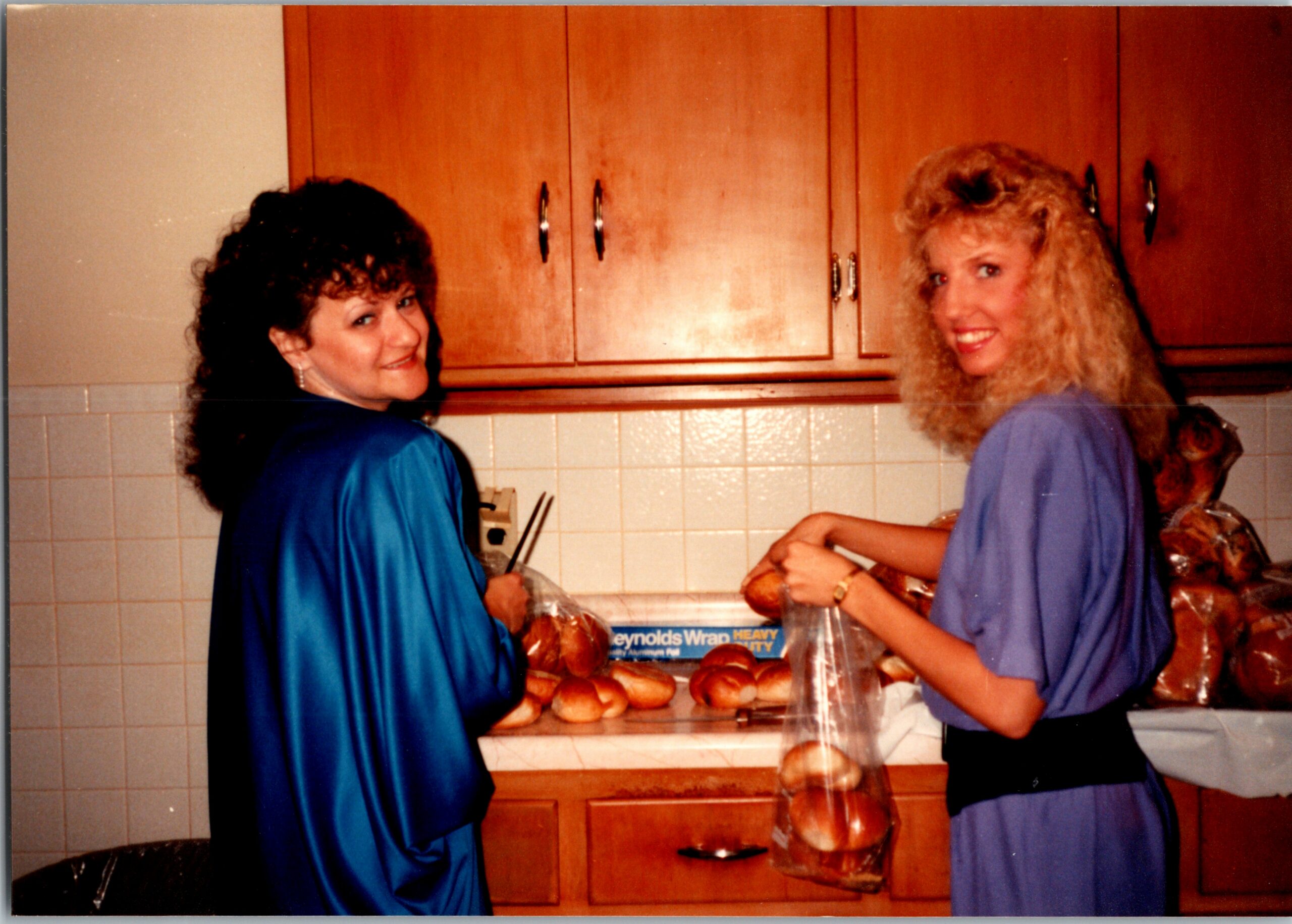 Two women in blue uniforms smiling while shaping bagels at a kitchen counter with a rack of bread nearby.