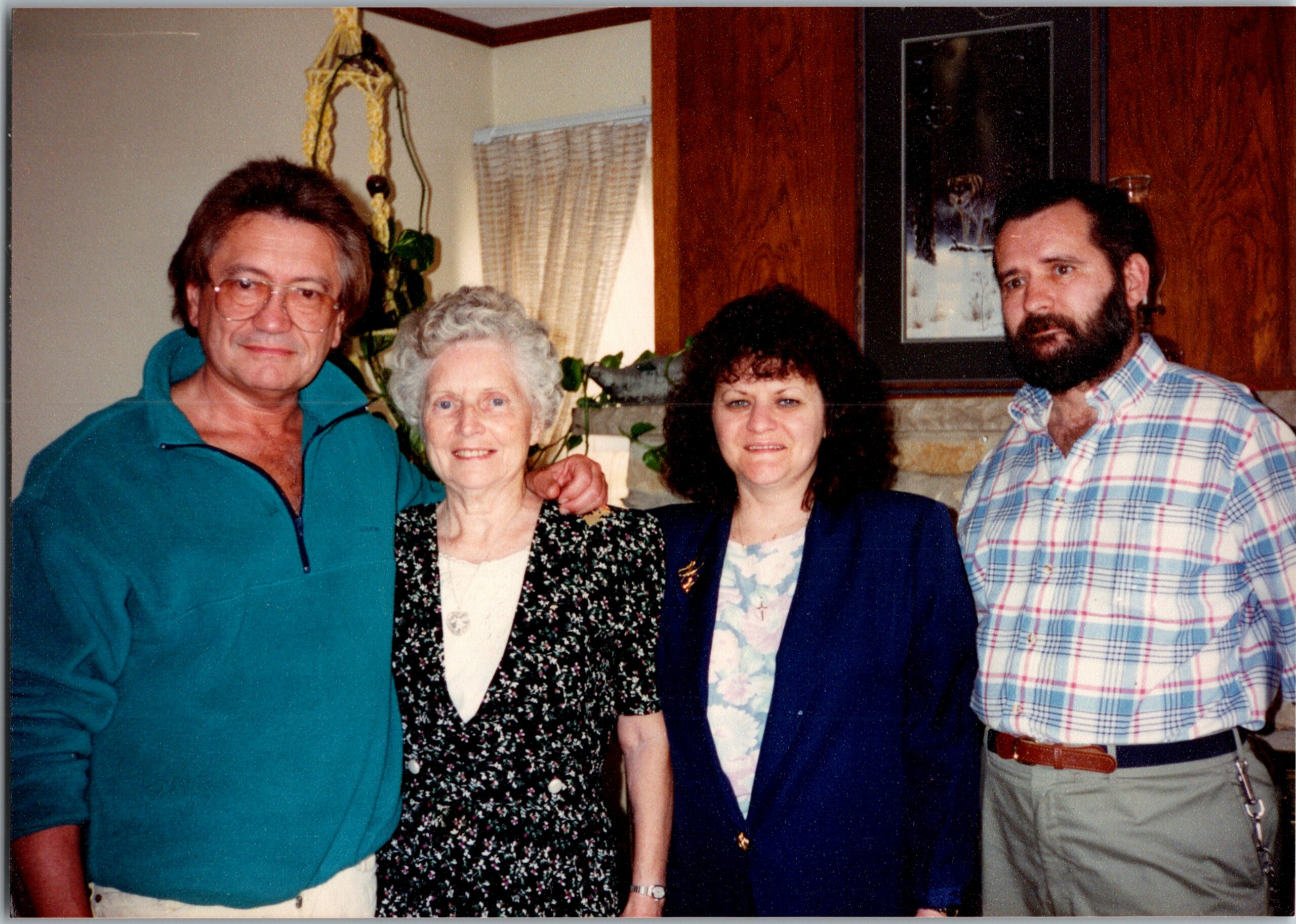 Group of four adults posing together indoors, near wood-paneled walls and a fireplace mantel. From left: man in teal zip-up sweater, elderly woman in floral dress, woman in a blue blazer, man with a plaid shirt and beard.
