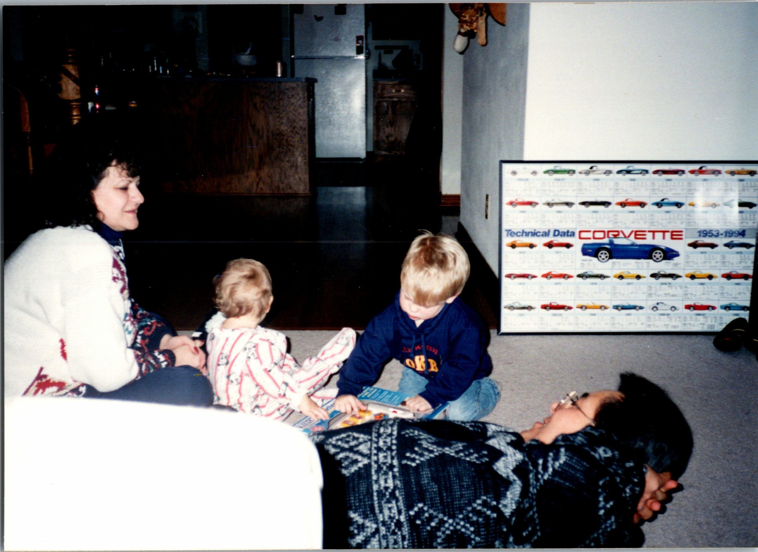 A woman with three small children sitting on the carpet, looking at a book together in a living room with a Corvette poster on the wall.