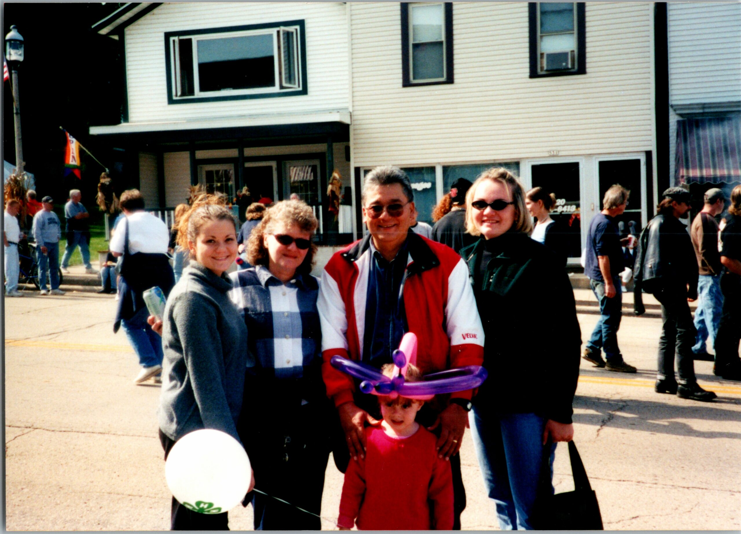 Group of four adults and a child posing for a photo on a sunny street with balloons, in front of a pale building.
