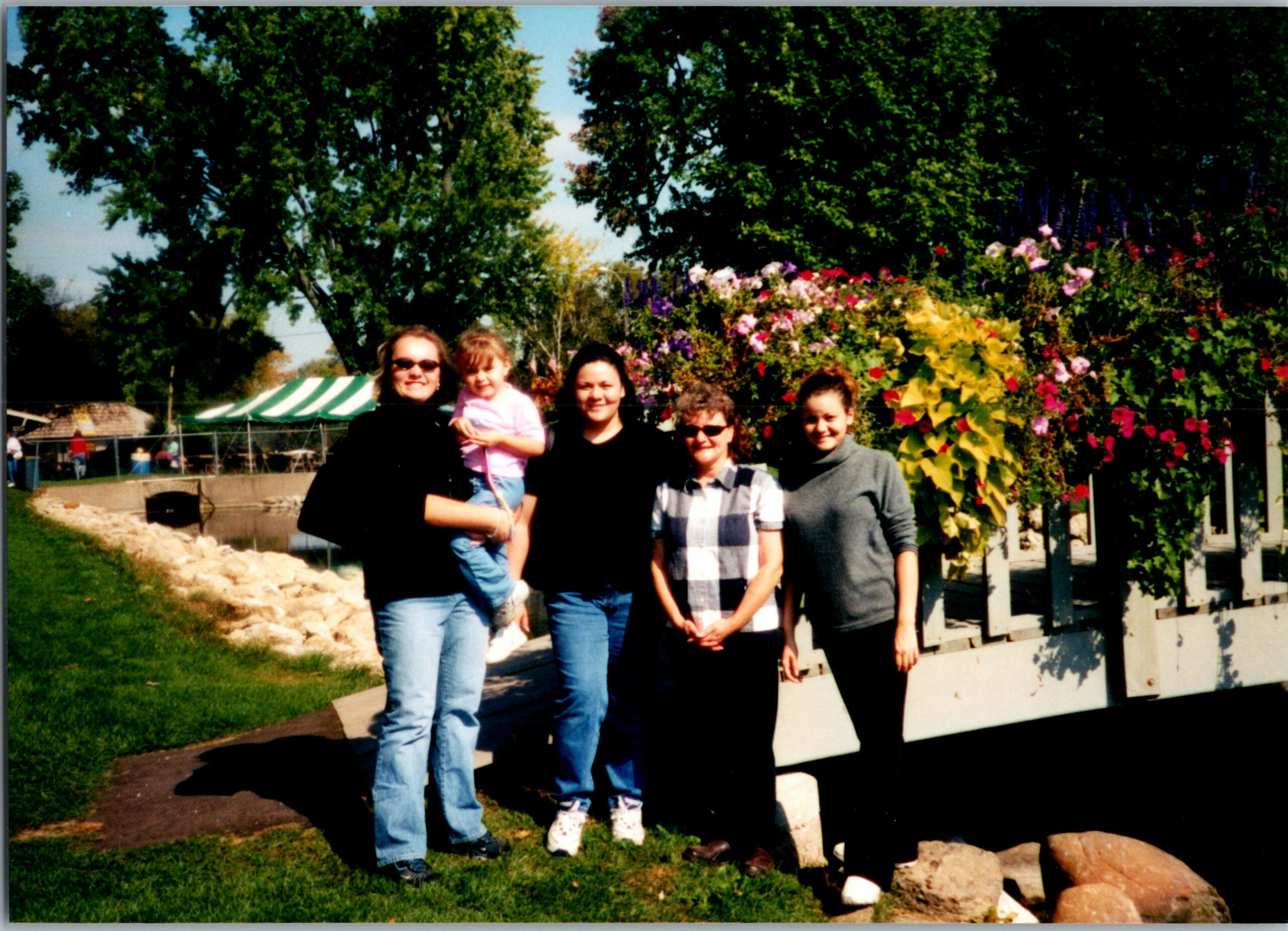 Group of people posing together outdoors in a park beside a colorful flower bed and a wooden fence on a sunny day.