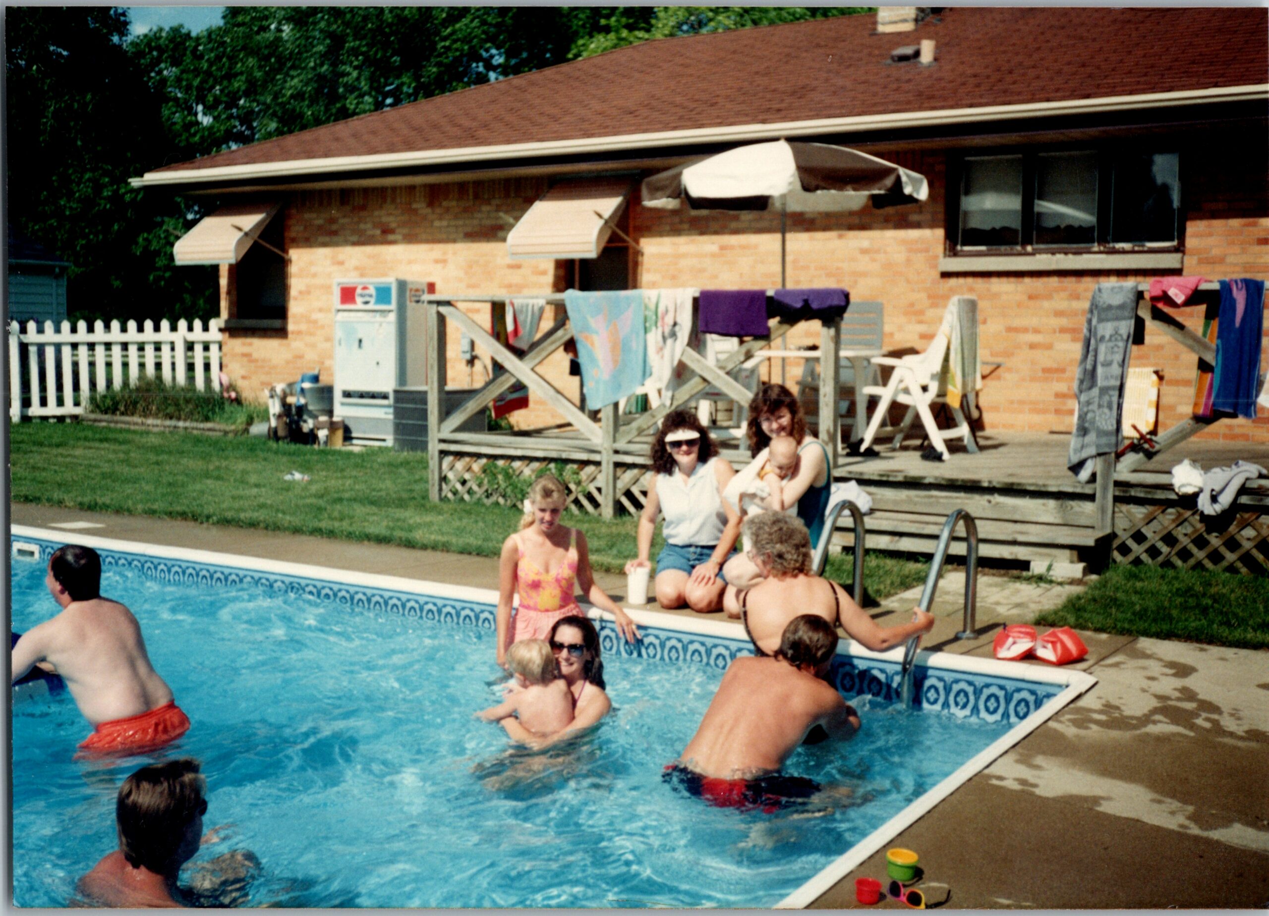 Family enjoying a sunny backyard pool party with people swimming and lounging near a wooden deck and brick house in the background, towels hanging on a fence rail.