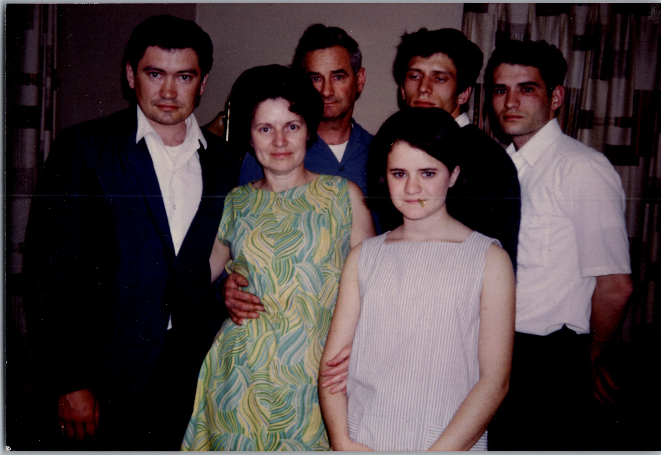 Six adults posing closely for a group photo indoors, with two women in front wearing patterned dresses.