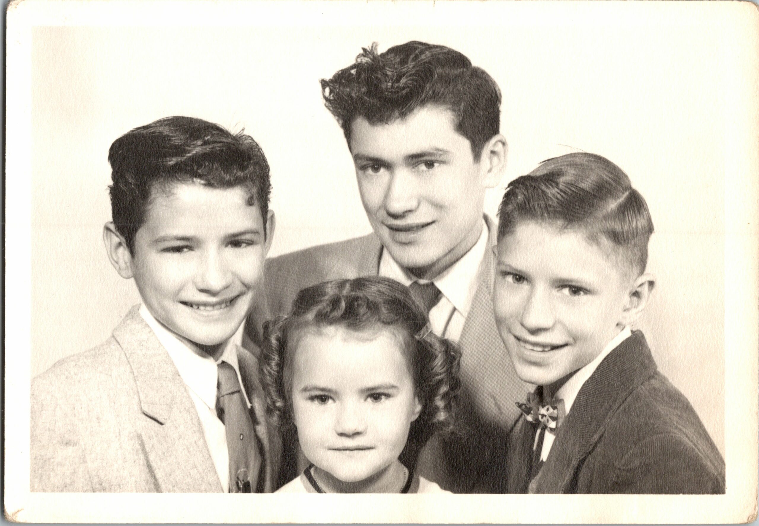 Group of five teens in formal attire posing closely for a vintage portrait, smiling at the camera.