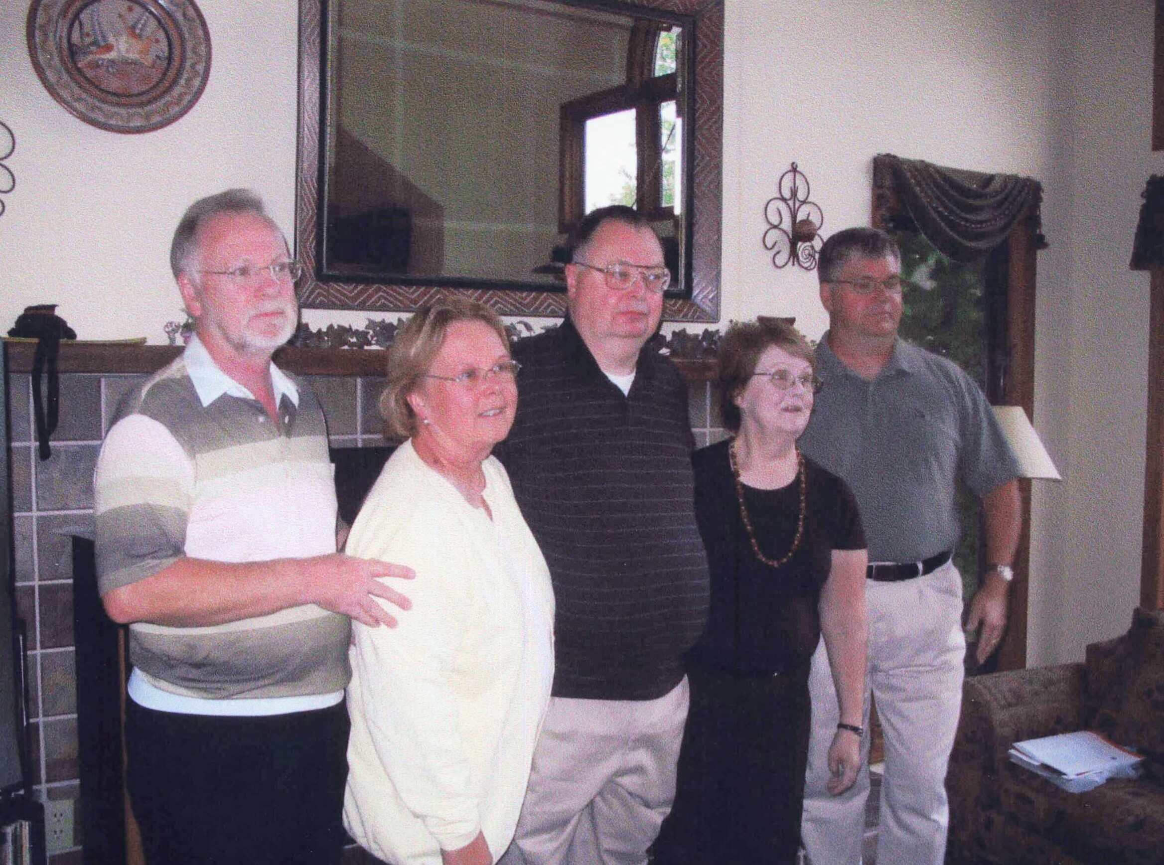 Five adults stand together indoors, posing for a group photo in a living room setting with a mirror and decor behind them.