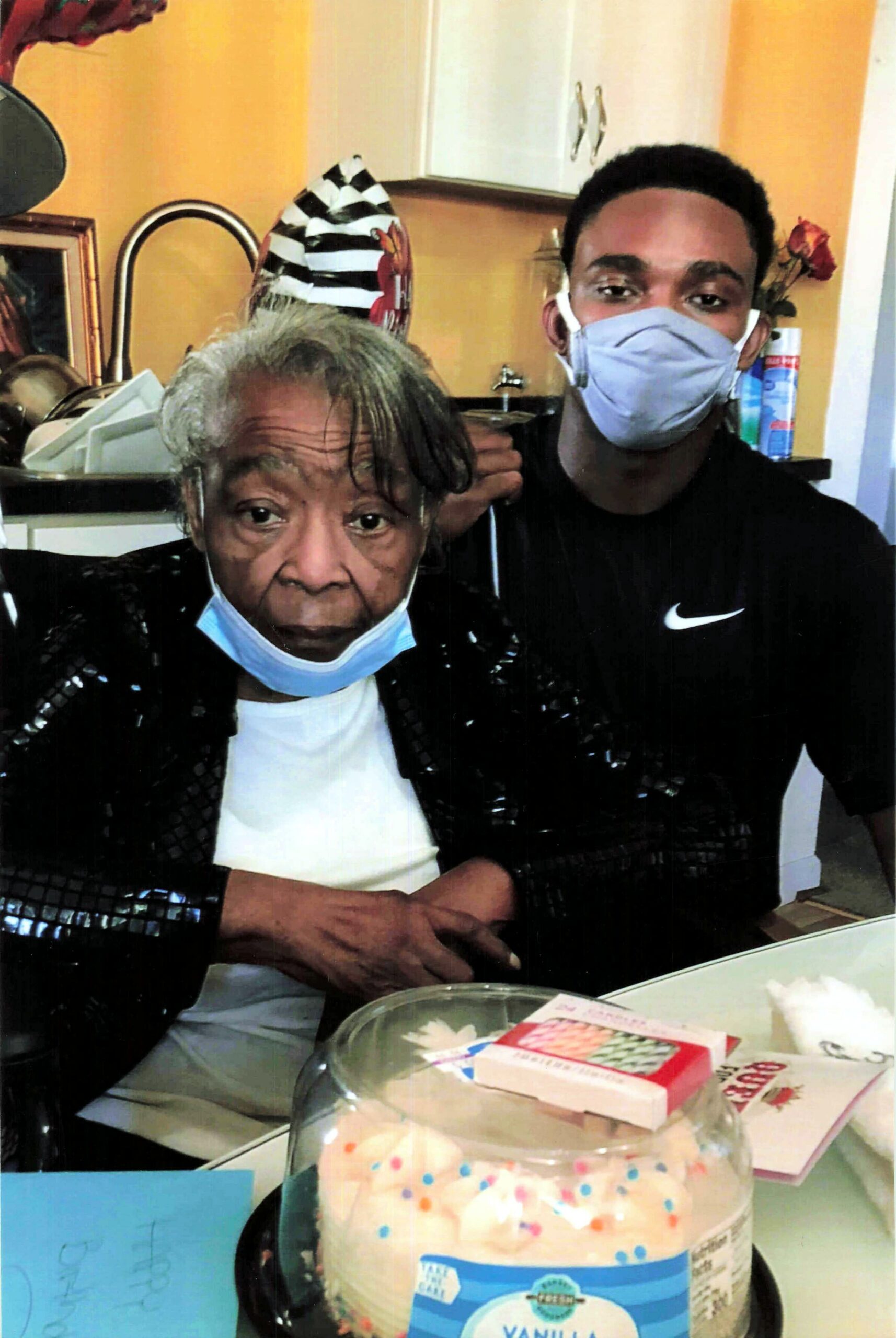 Older woman and younger man wearing masks sit together at a kitchen table with a sprinkled birthday cake under a glass dome in the foreground.