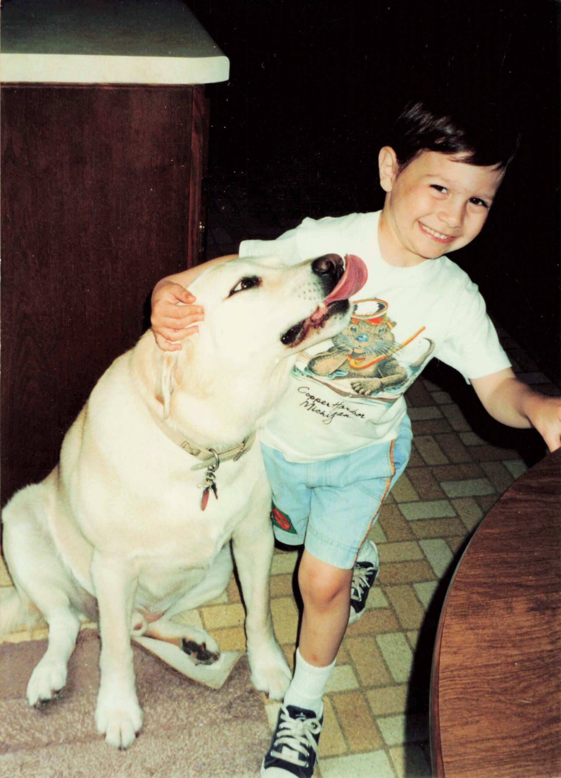Smiling boy in a white shirt petting a large white dog with its tongue out indoors.