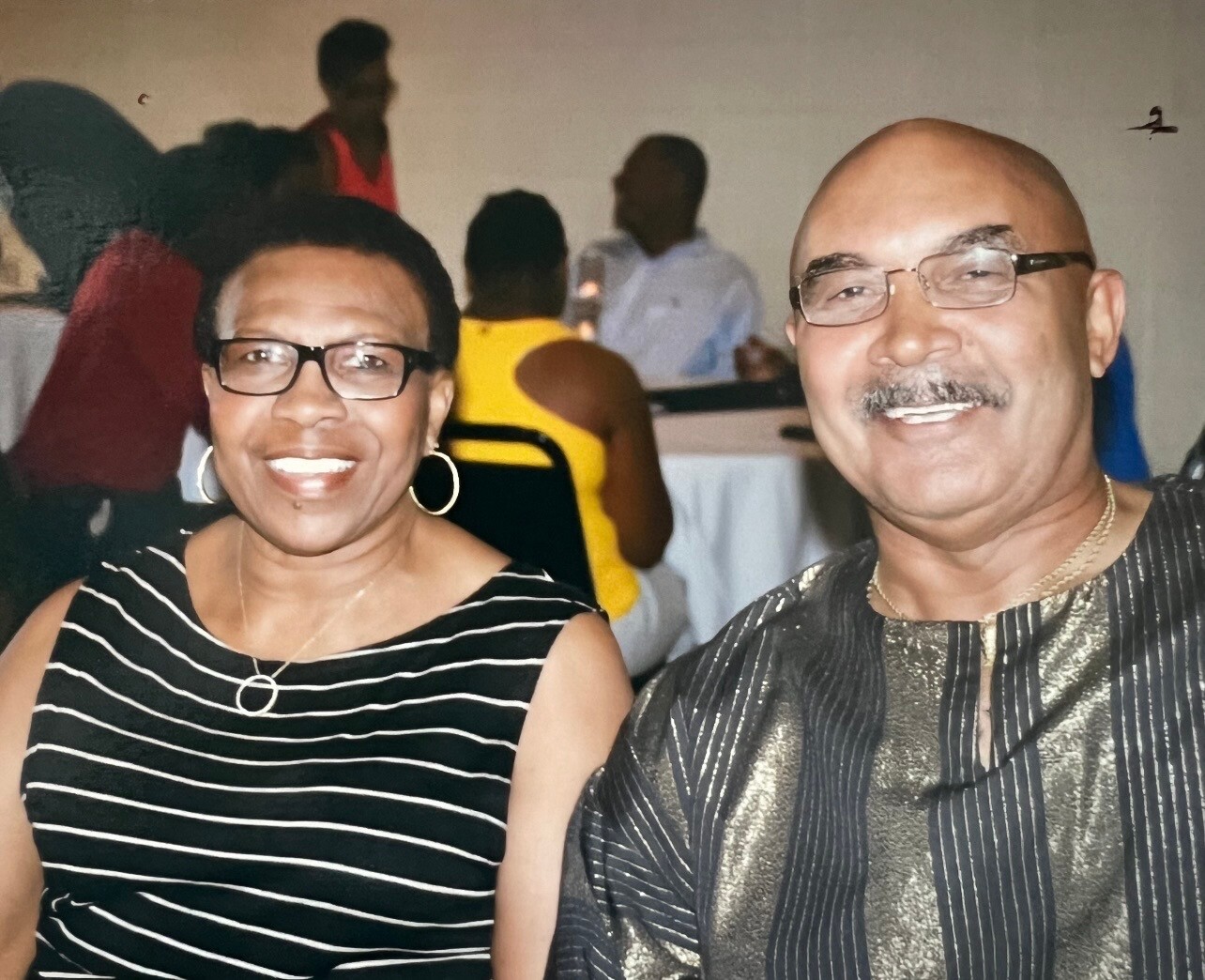 Smiling couple posing for a photo at a social gathering; woman in a black and white striped sleeveless dress, man in a metallic striped shirt and glasses.