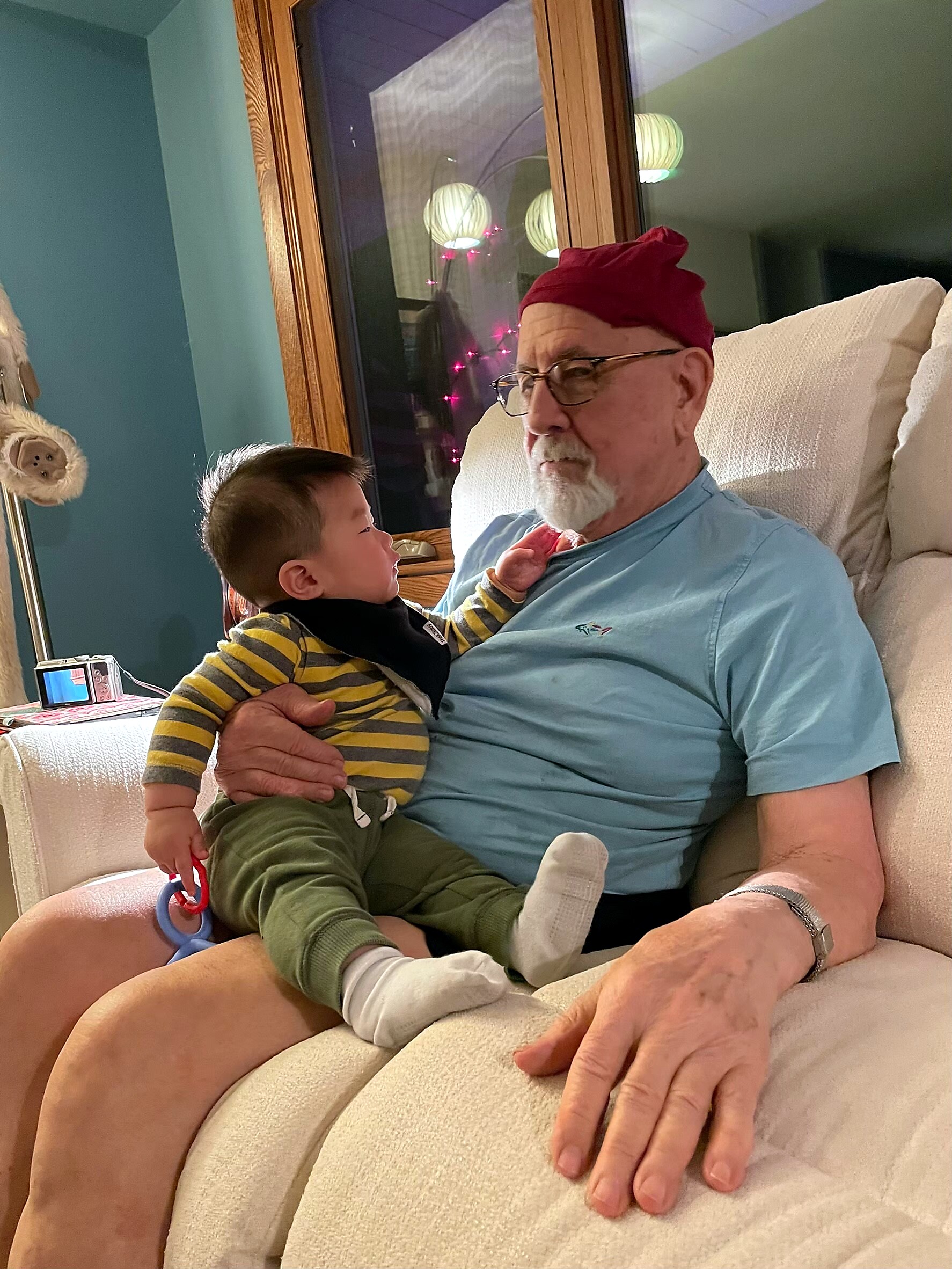 Grandfather in a red cap sits on a cream couch, holding a young child facing him.