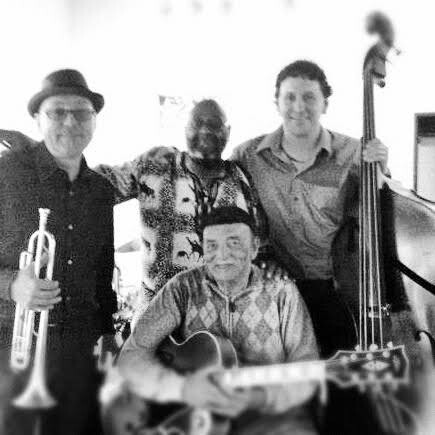Five male musicians posing for a photo in a room: trumpet player on the left, guitarist seated center, upright bass player on the right, and two others smiling in back.