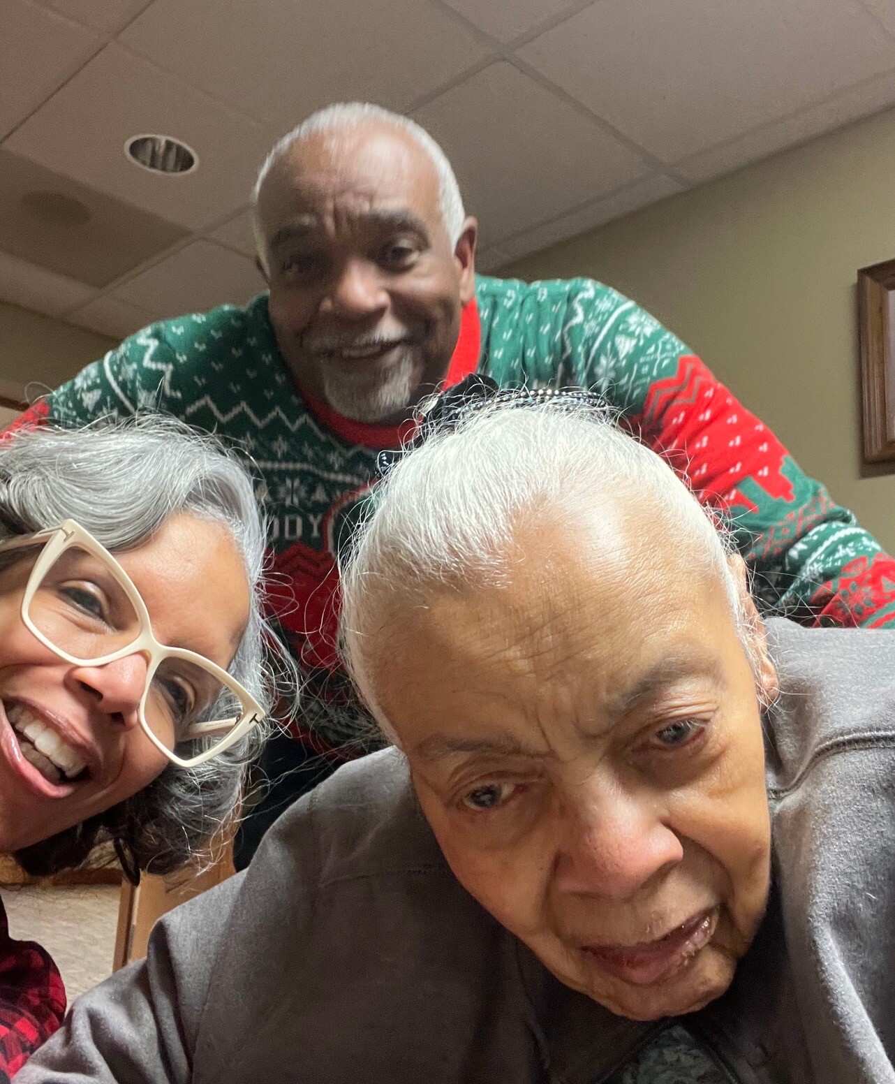 Three seniors take a cheerful selfie indoors: a woman with white glasses on the left, a man in a green holiday sweater in back, and another elderly person in the foreground smiling.