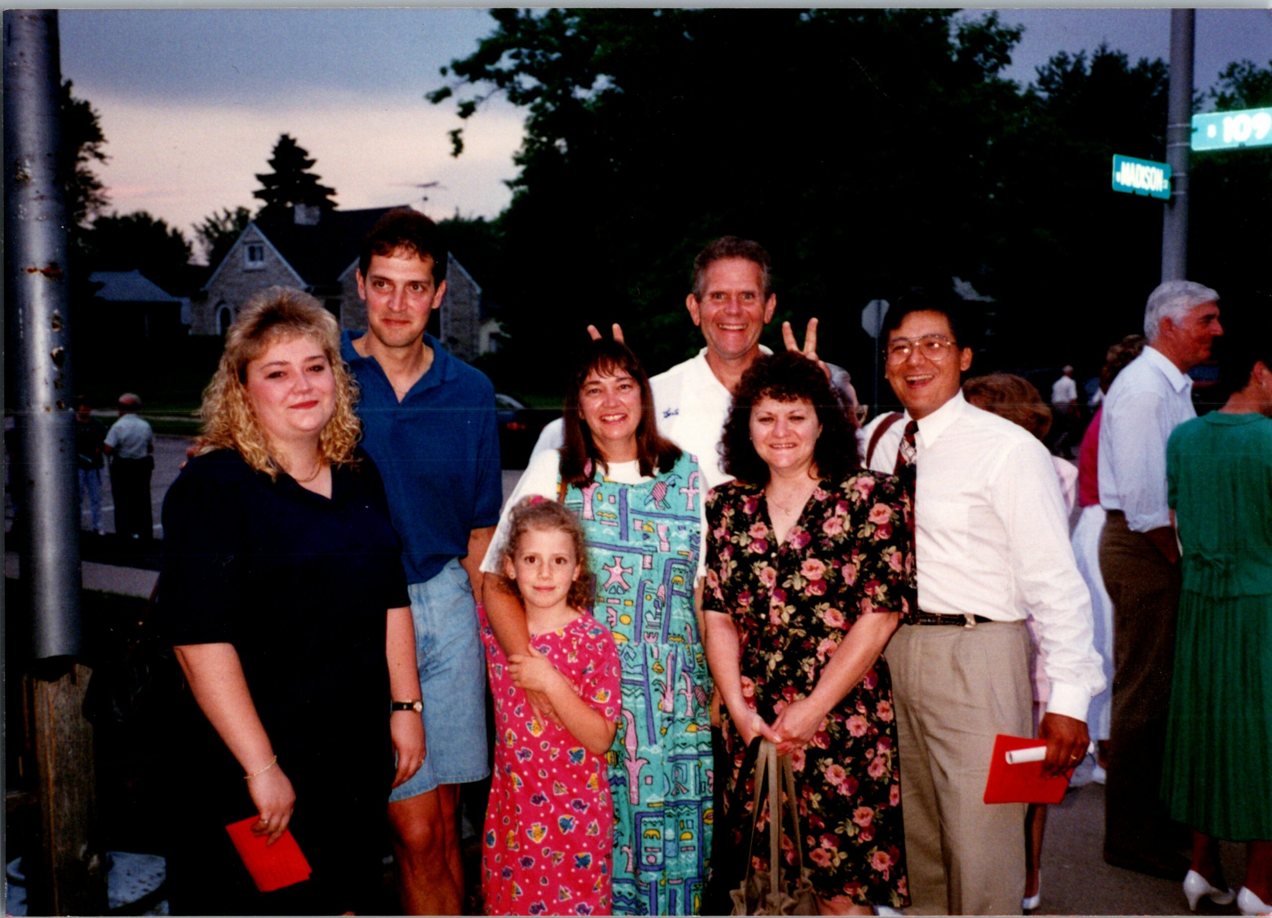 Group of adults and children posing for a photo on a street corner, smiling at the camera, with houses in the background.
