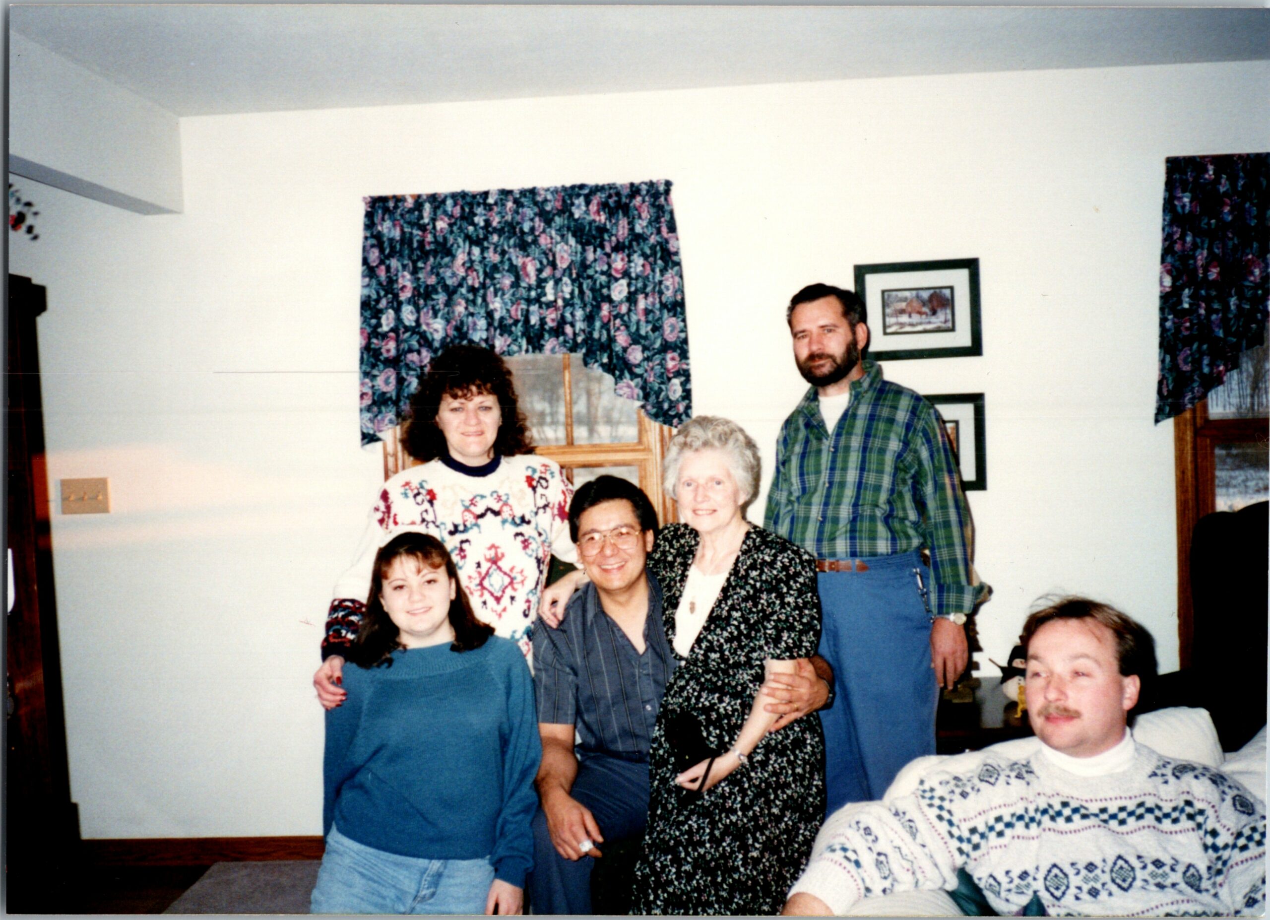 A group of people posing indoors for a family photo in a living room with floral curtains and framed art on the wall.