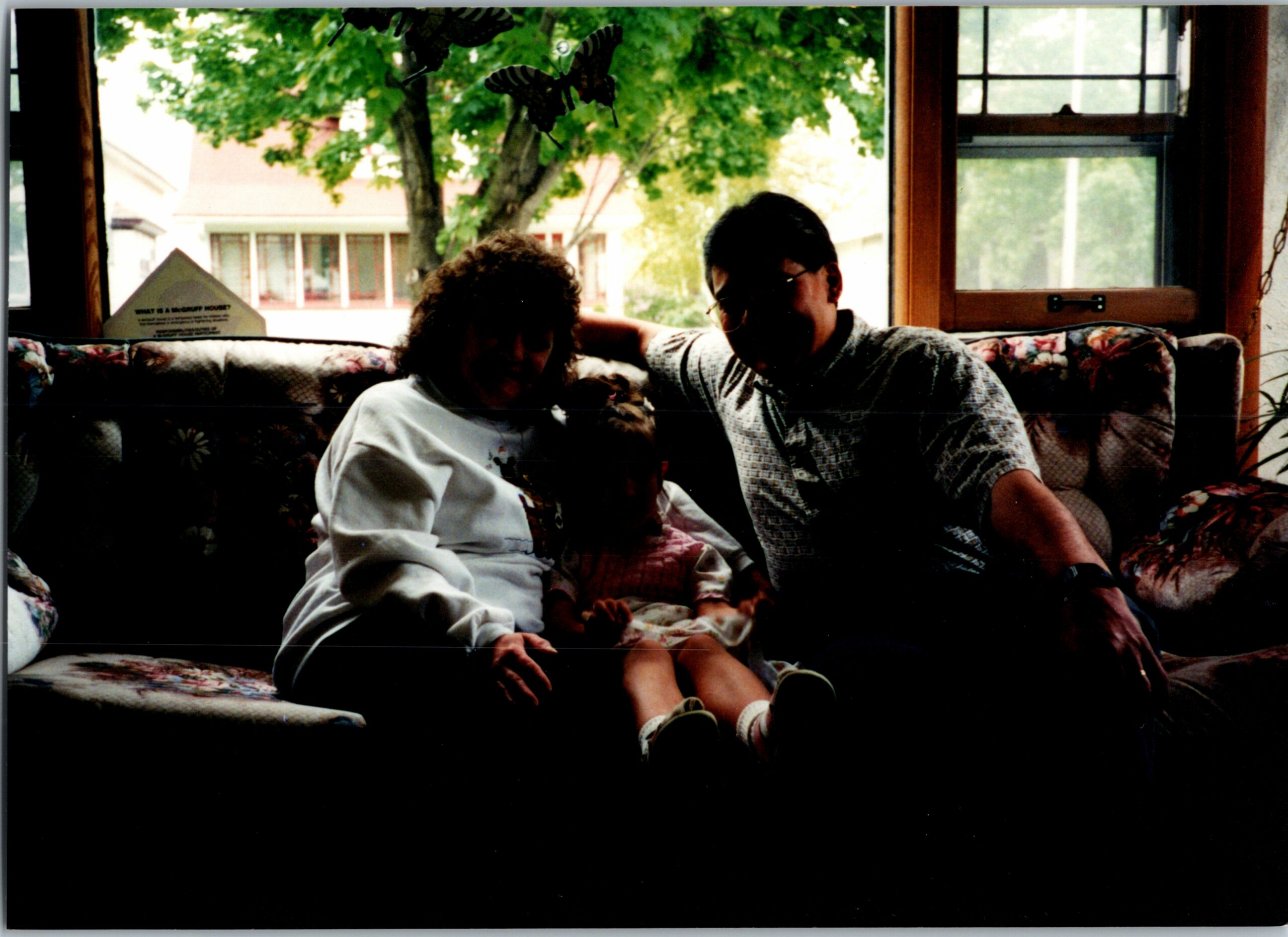 Two people sit on a floral sofa in a bright living room, with a tree visible through the window behind them, and a small dog on the laps of the children.