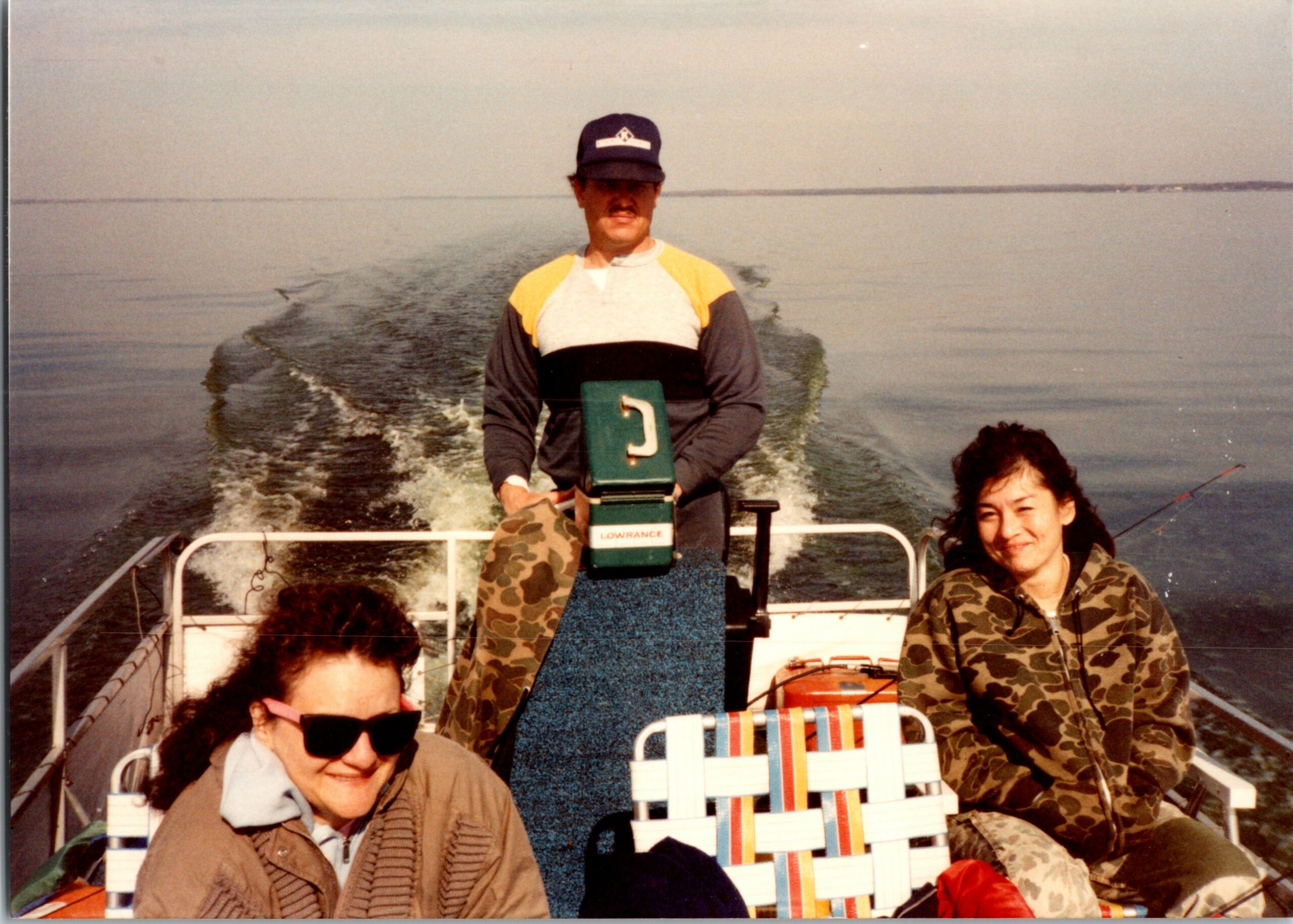 People riding in a small boat on calm water; a man standing in the center wears a navy cap and camouflage clothing, holding a green Lowrance fish-finder device while others sit nearby.