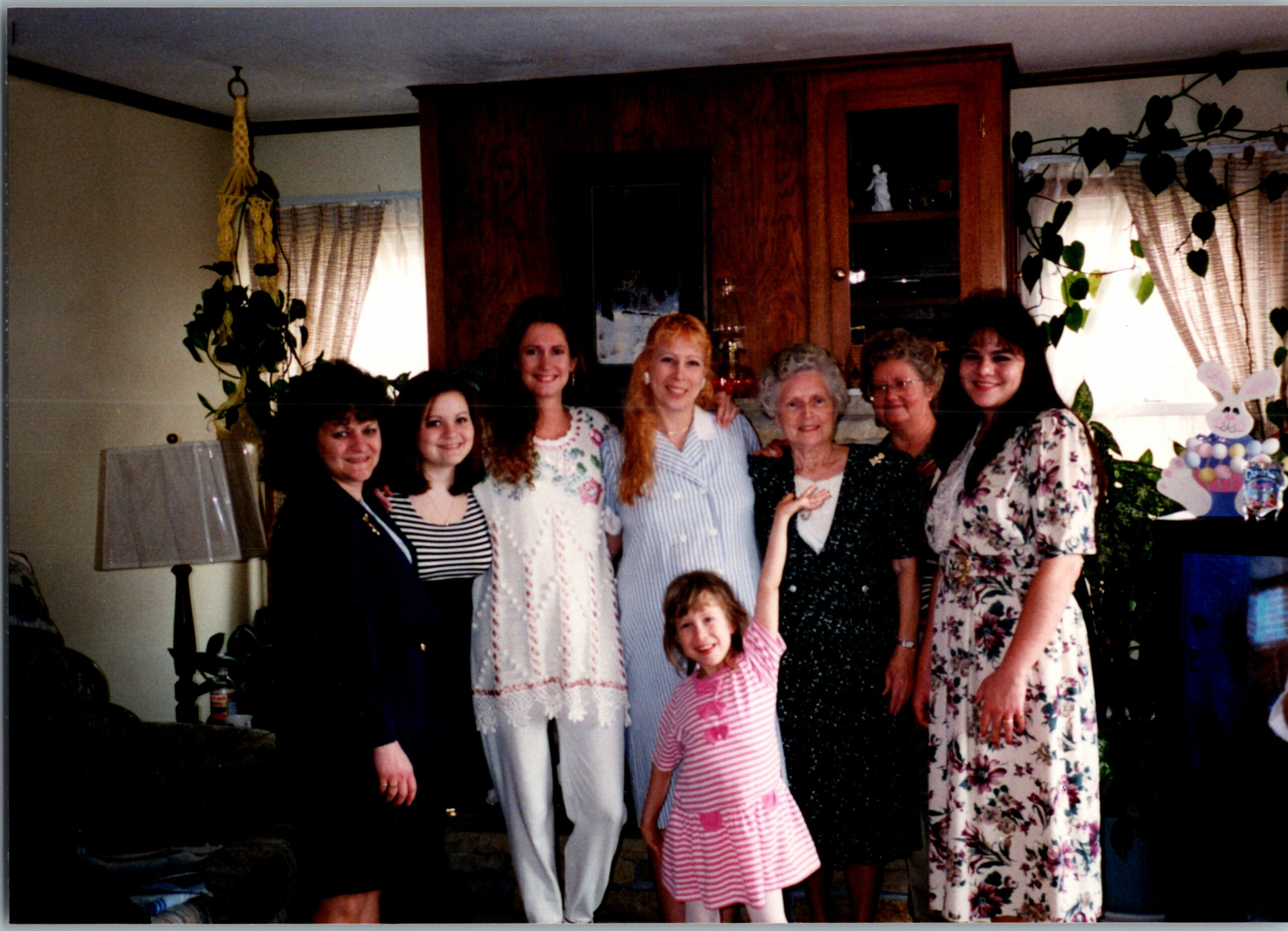 Group of nine people posing in a living room, smiling, with plants and a bunny-themed cake visible on the right side of the room.