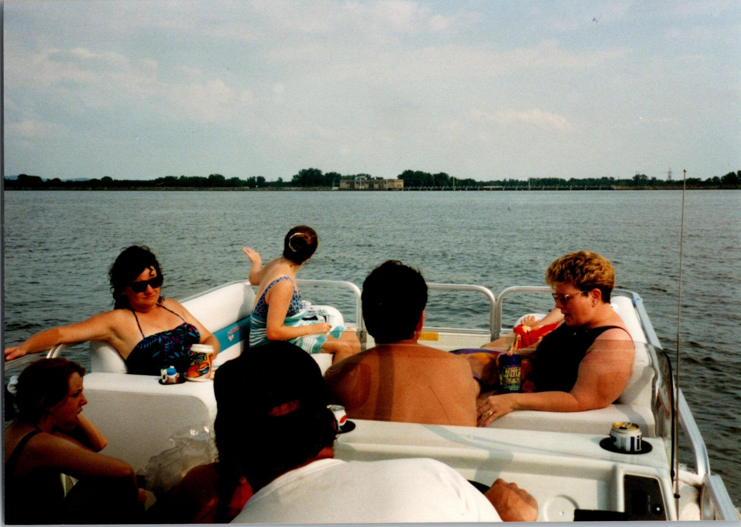 People relax on a small motorboat with drinks, sunbathing against a lake shoreline under a blue sky.