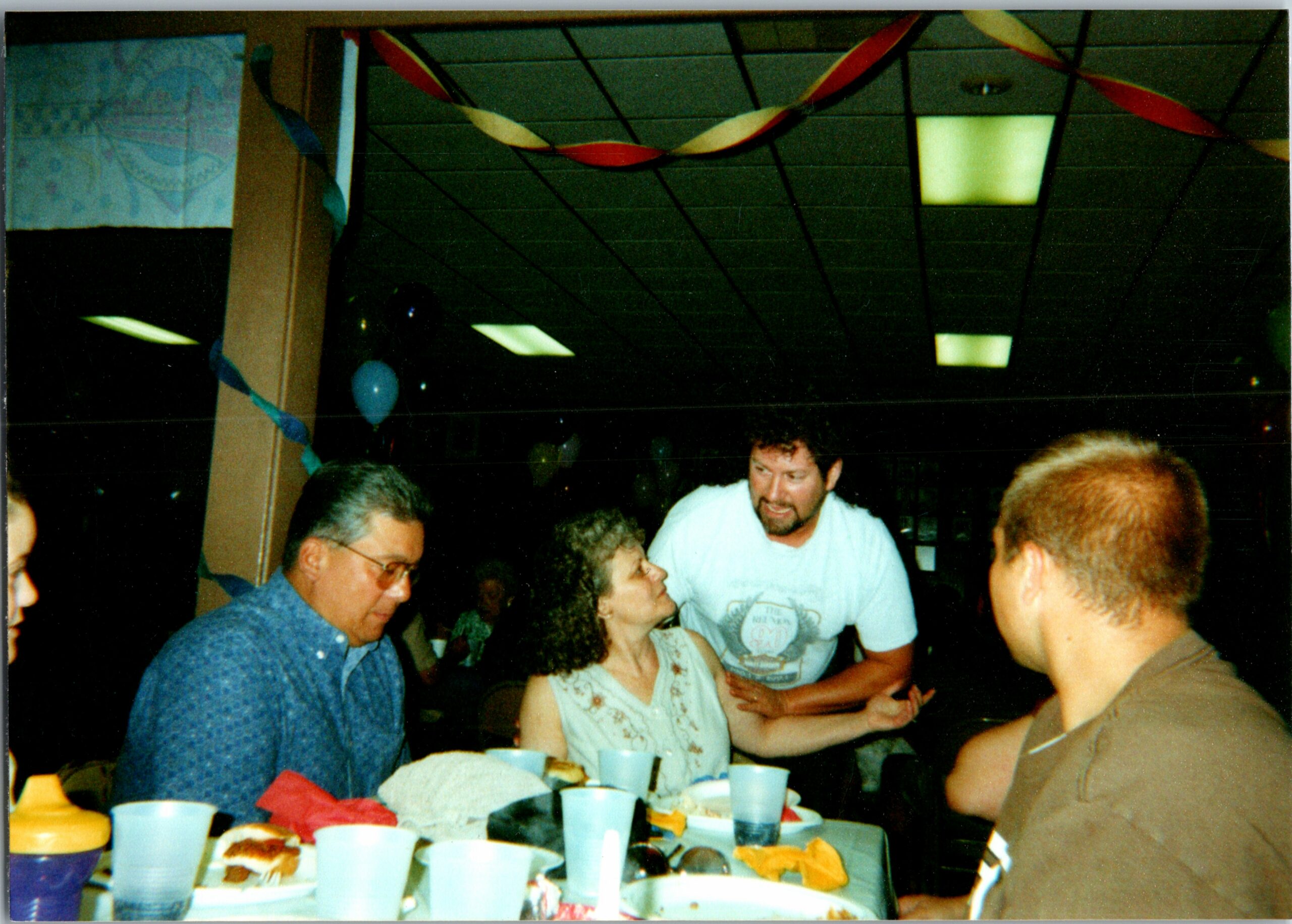 People gathered around a table with snacks and drinks at a party, colorful streamers overhead in a dimly lit room.