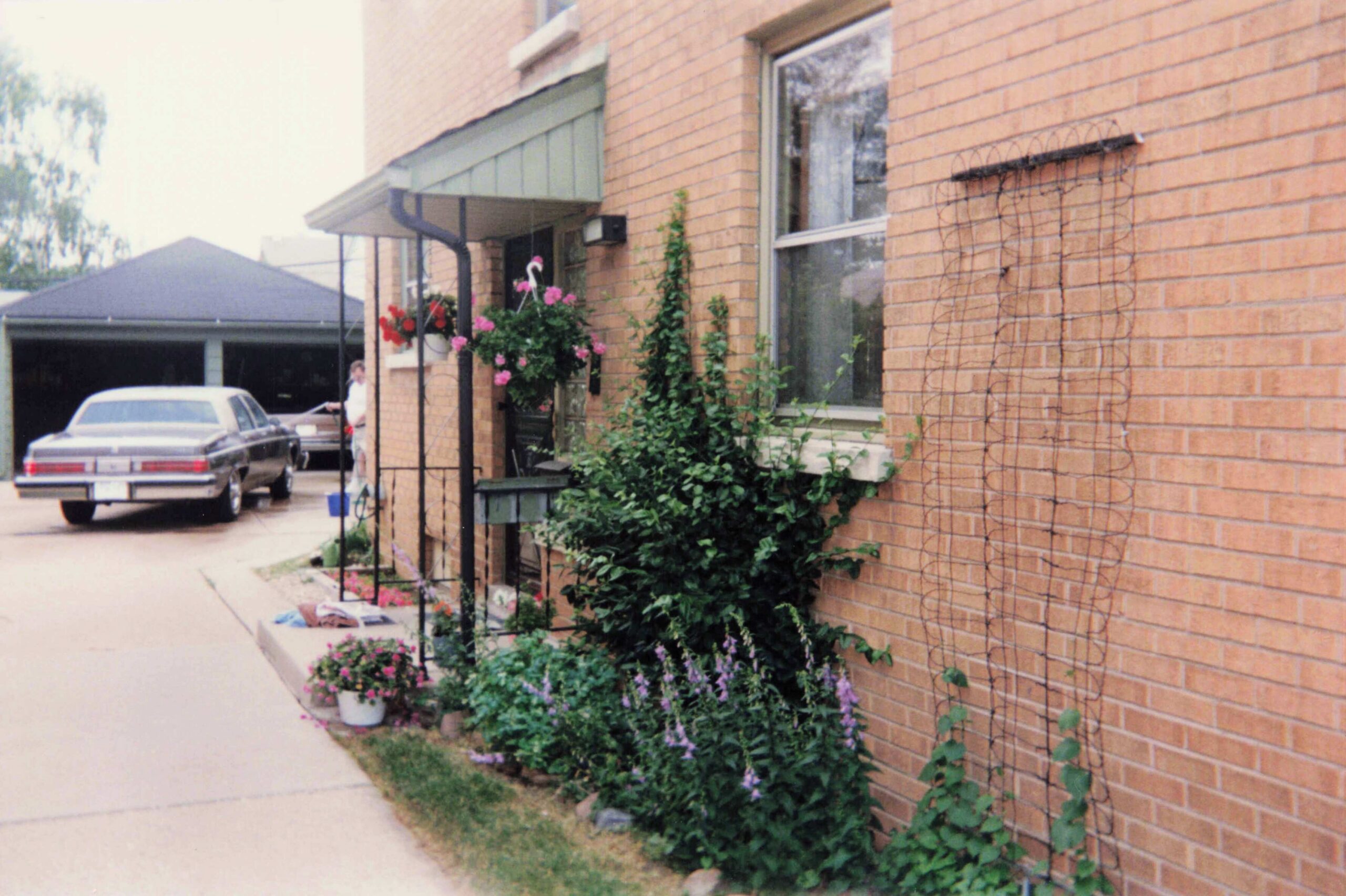 Brick townhouse with flowering planters along the walkway, a vine on the wall, and two cars in the driveway.