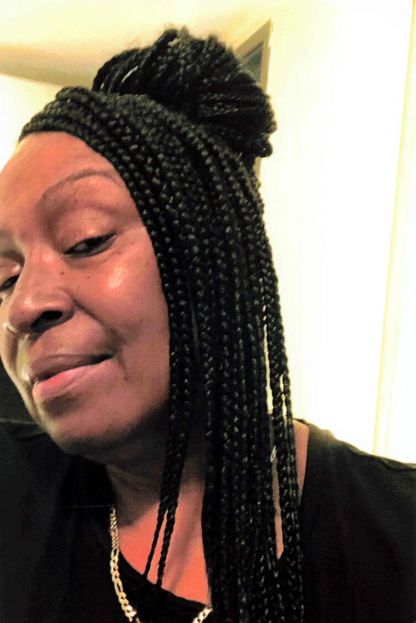 Partial selfie of a person with long black box braids, wearing a gold chain and a black top, smiling slightly at the camera.