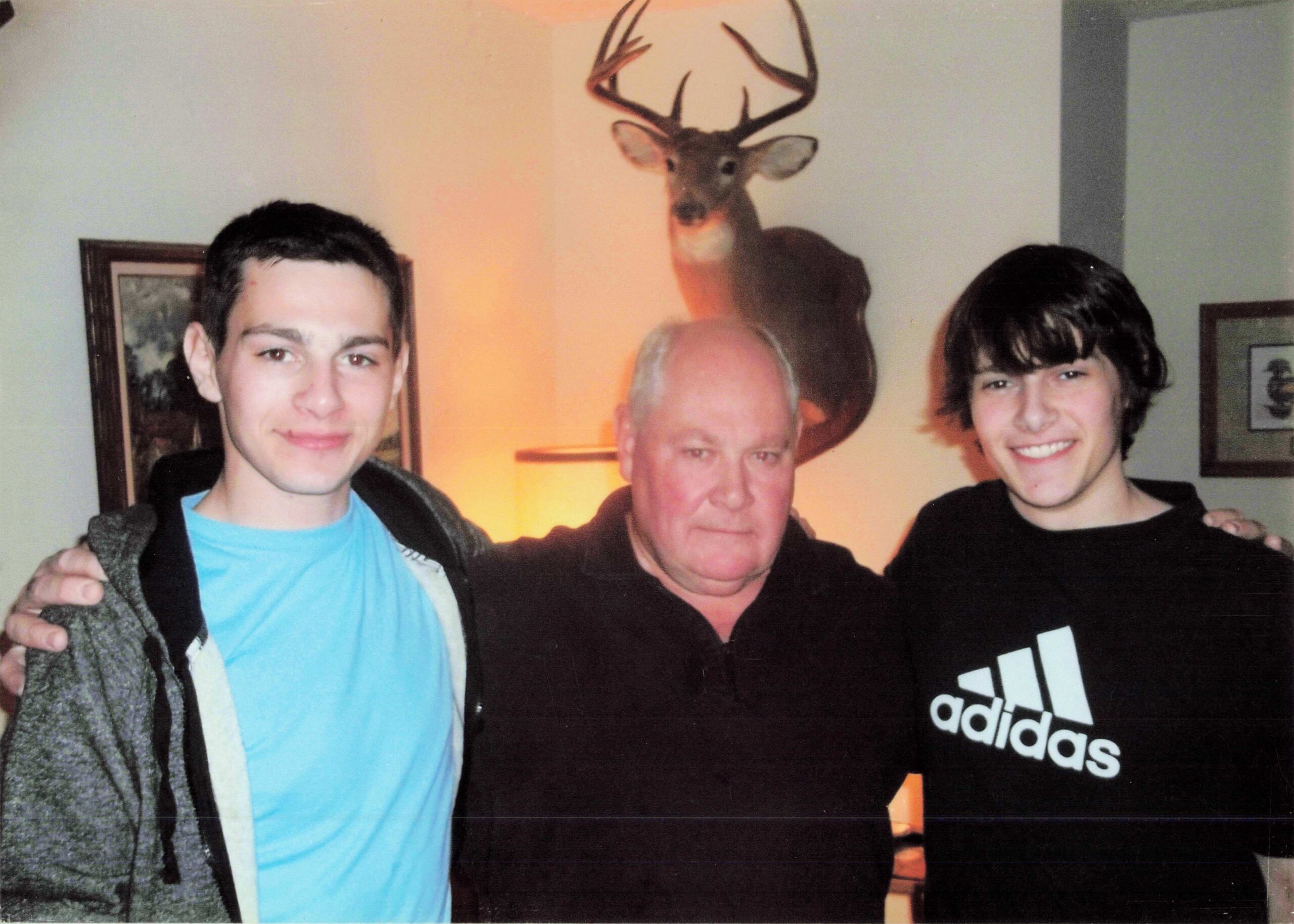 Three men pose with arms around each other in a room, a mounted deer head is on the wall behind them.