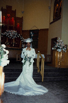 Bride in a white wedding gown and veil holding a bouquet, standing at the church aisle with floral arrangements visible in the background.