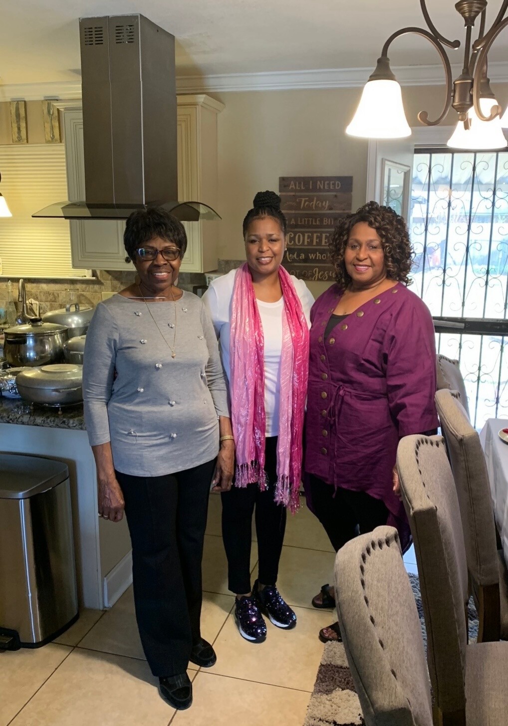 Three women stand together in a home kitchen and dining area, smiling for the photo, with pots on the stove and a chandelier overhead.
