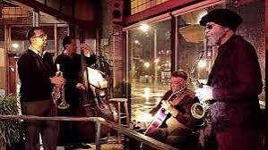 Nighttime cafe scene with a small jazz trio—saxophonist, clarinetist, and accordionist—performing for a few onlookers.
