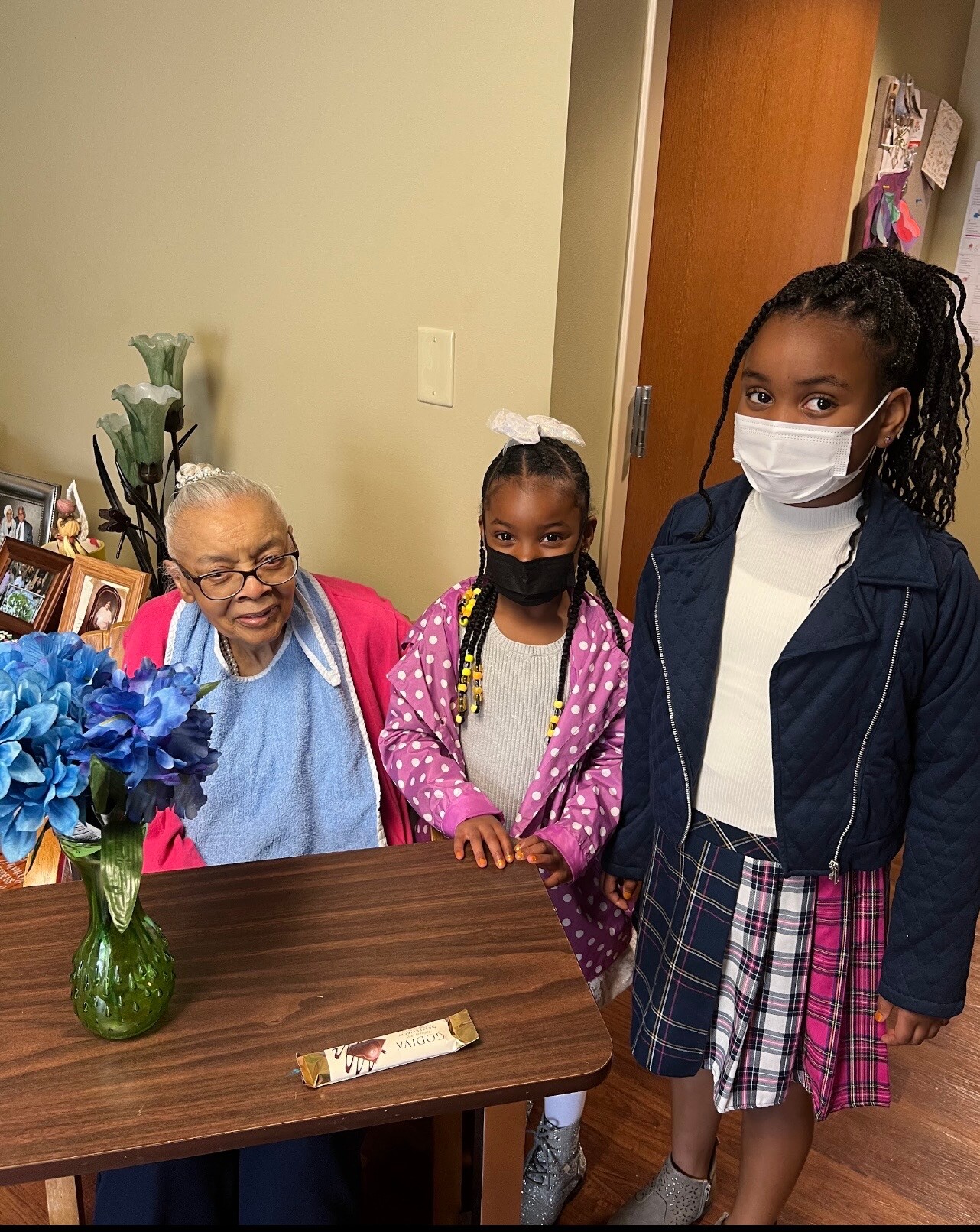Three people pose together in a room: an elderly woman in a pink cardigan sits at a wooden table with two masked girls standing beside her; a blue hydrangea bouquet in a green vase sits at the front of the table.