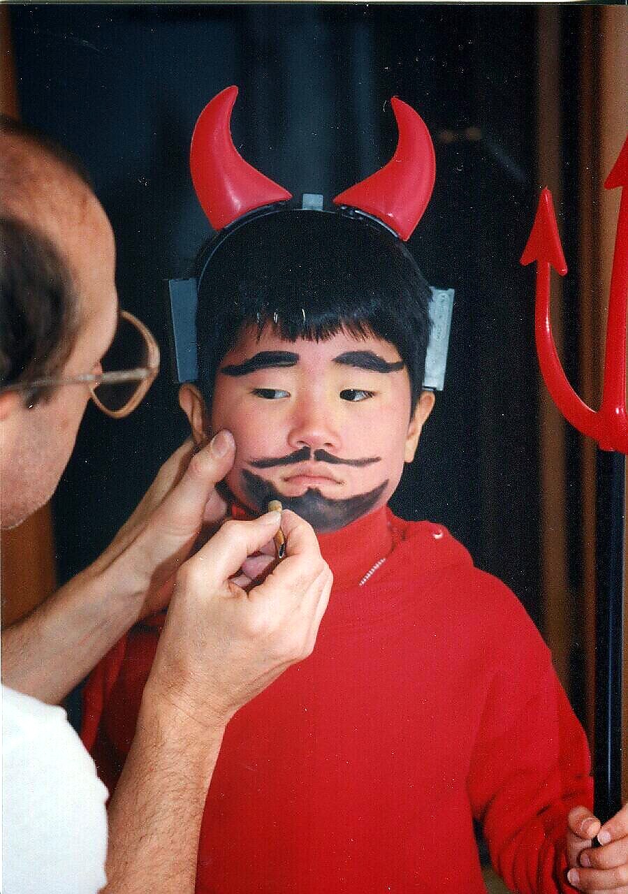 Child in a red devil costume has his face painted with a black mustache and beard by an adult painter.