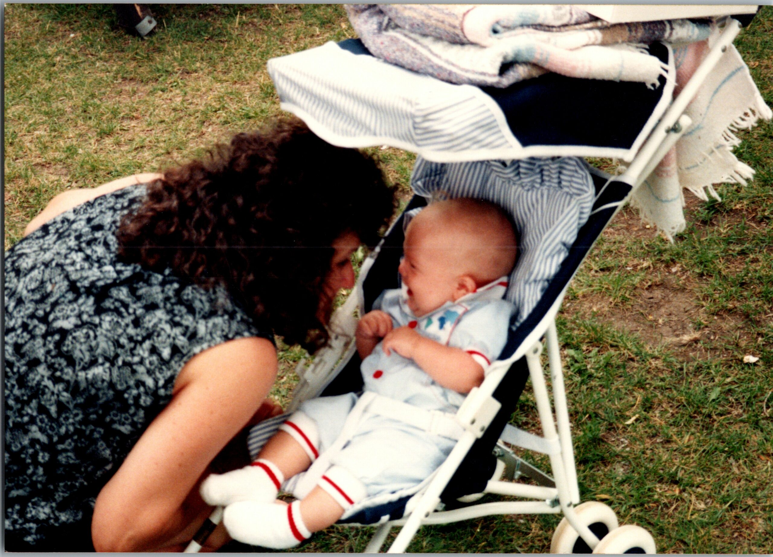 Woman bends toward a baby in a white stroller outdoors on grass, eyeing the infant.