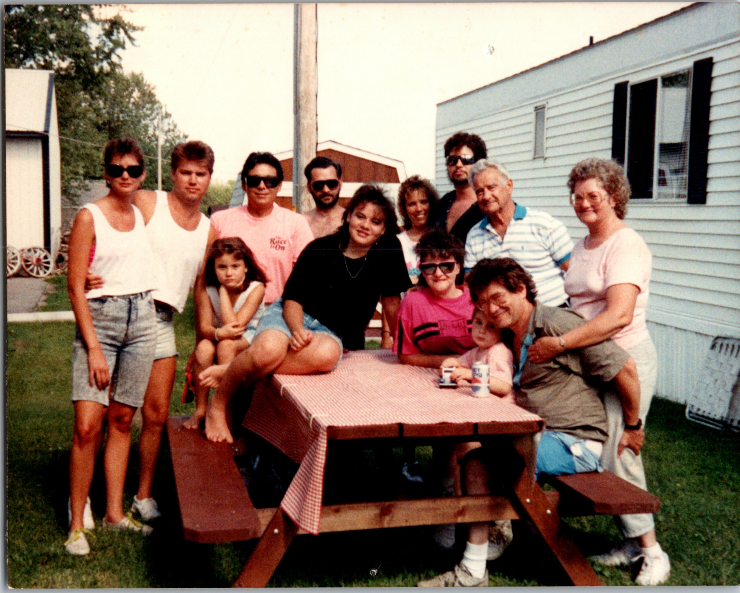 Group of people posing around a red-checkered picnic table outside a mobile home on a sunny day.