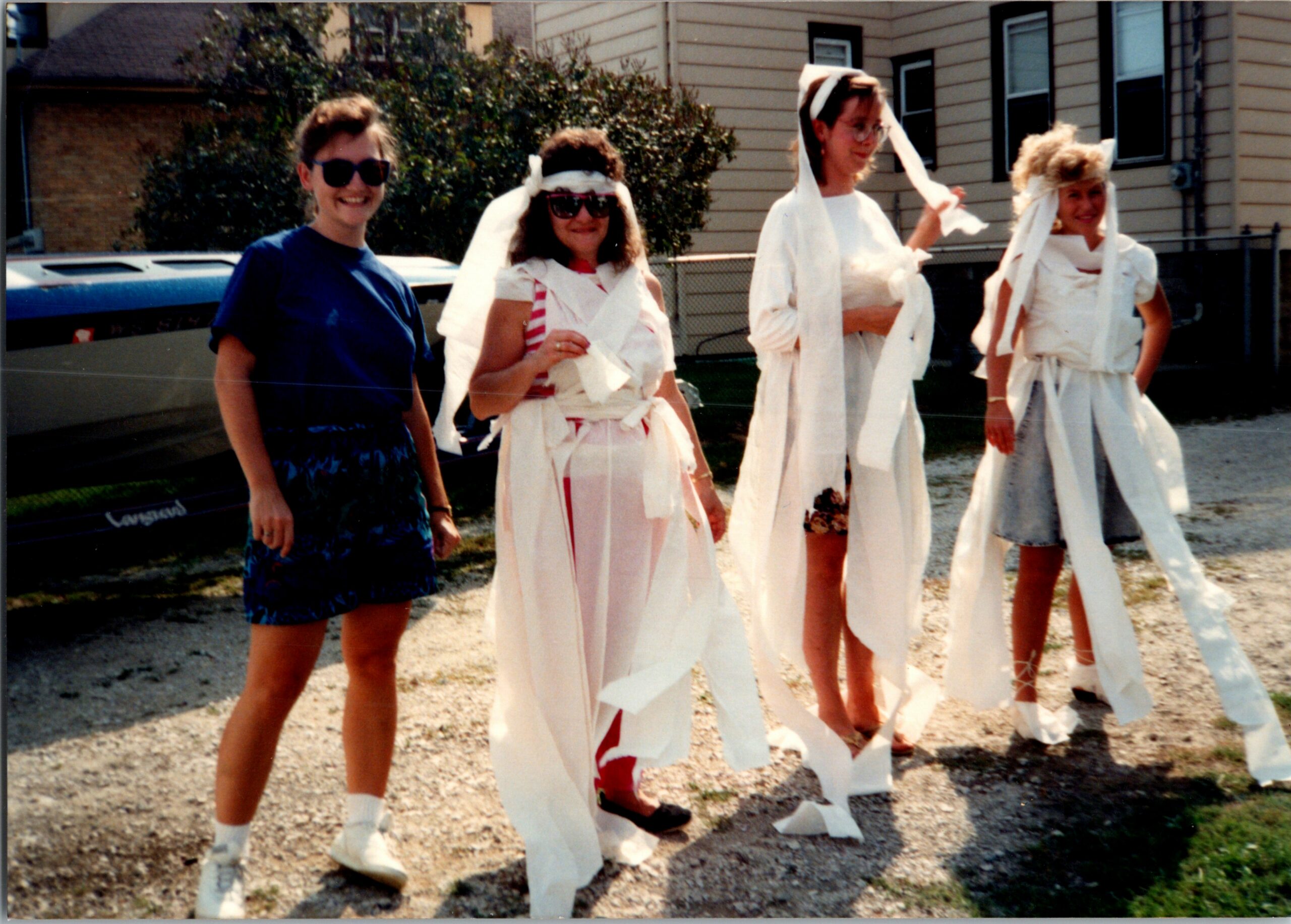 Four women stand outside, wrapped in long white streamers as if part of a playful costume, smiling for the camera.