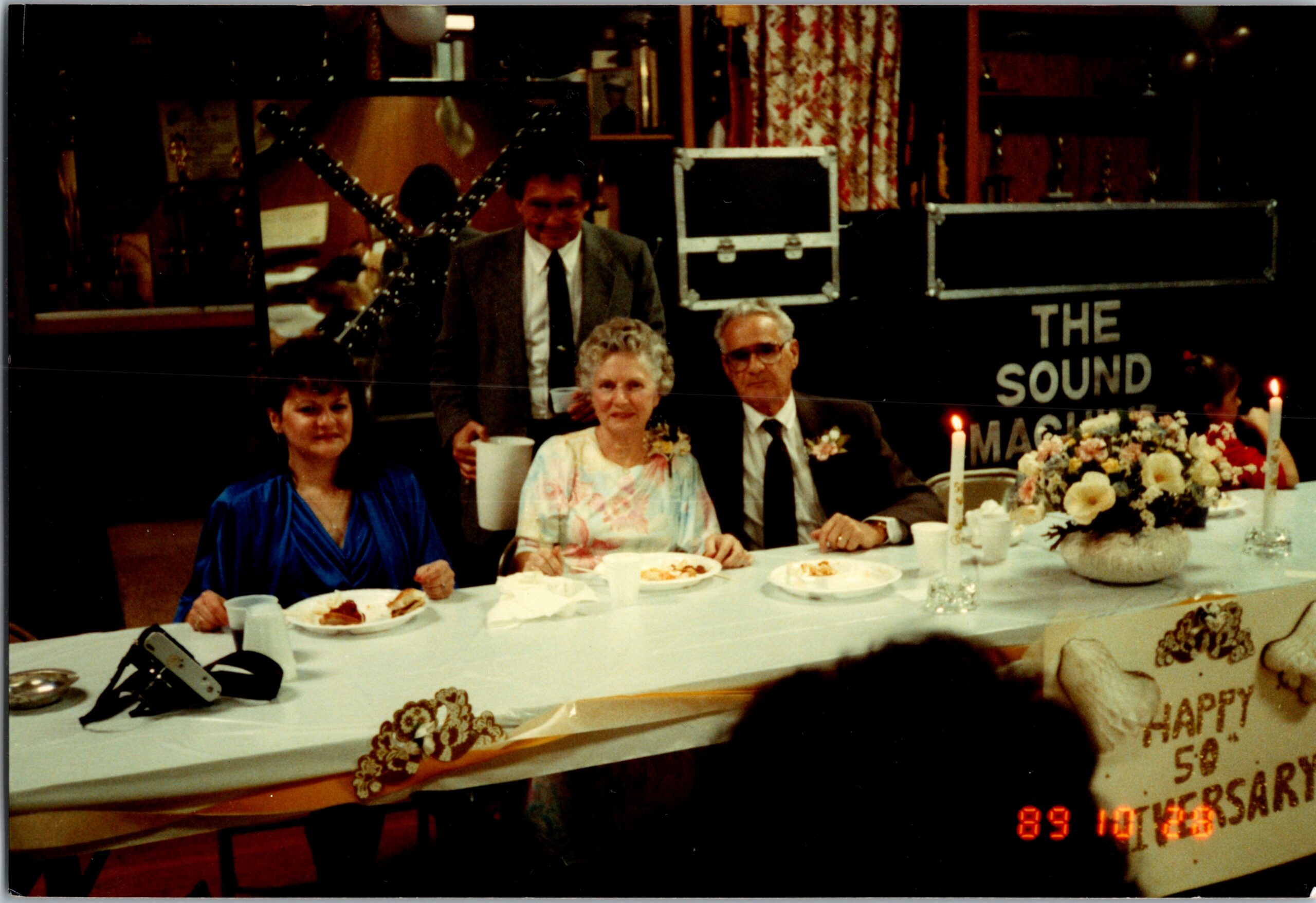 Four adults seated at a long birthday/party table with plates and flowers, posing for a photo, date stamp visible in the corner.